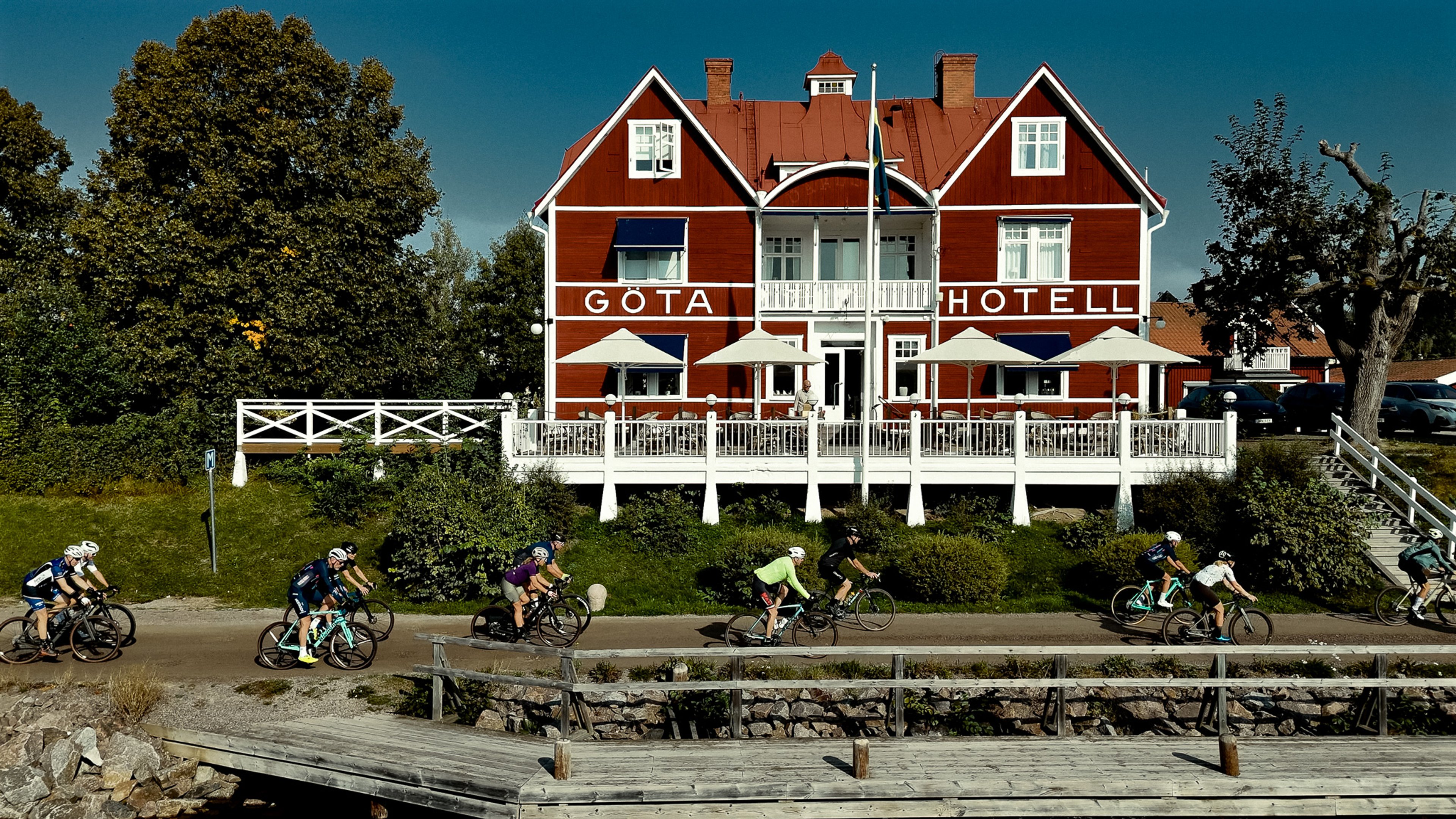 A group of cyclists rides along a path in front of the red Göta Hotell.