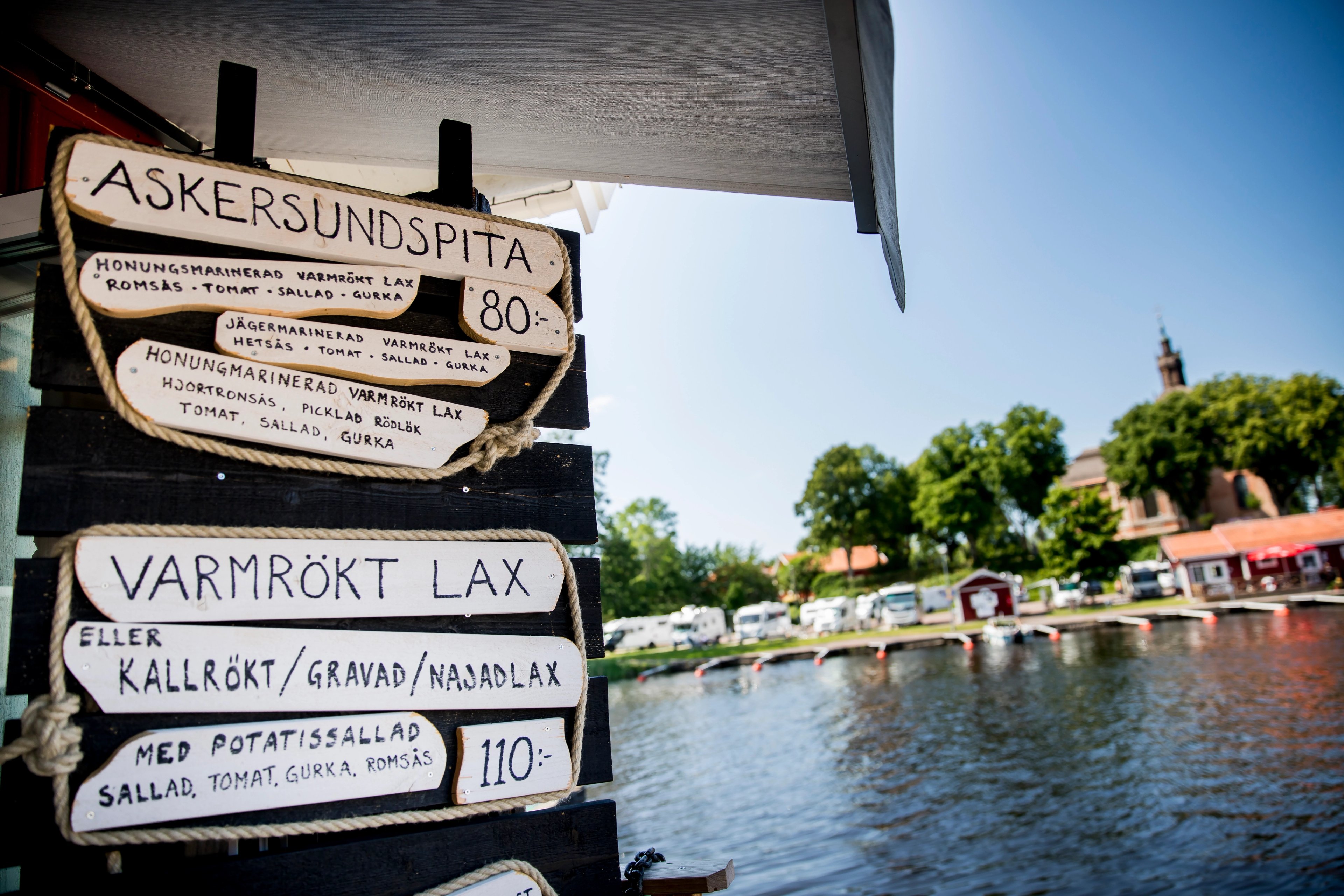 A wooden menu sign advertising Swedish salmon dishes for 80 and 110 SEK, with a blurred sunny waterfront background featuring trees and buildings.