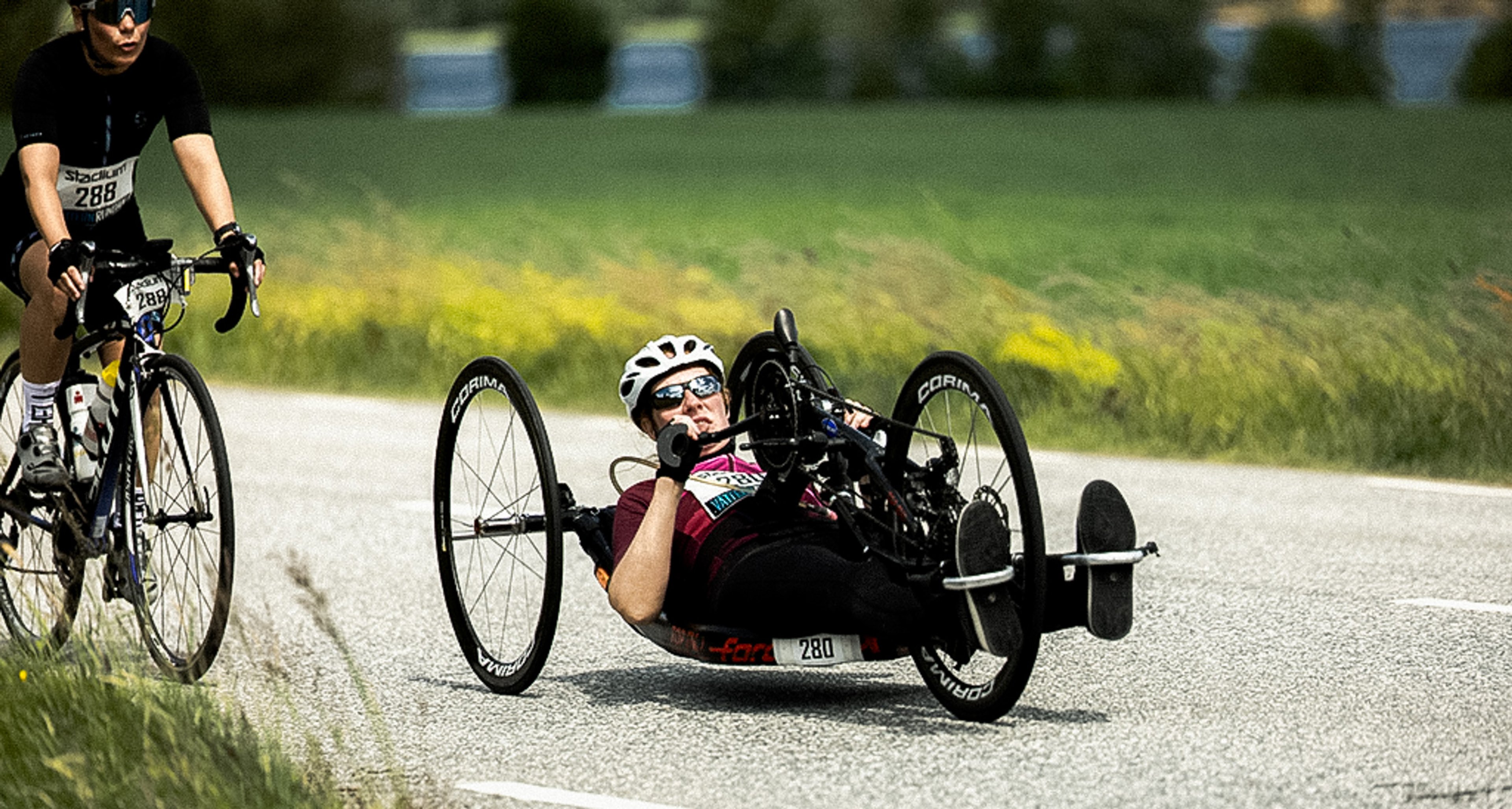 A person on a road bike and a person on a recumbent handcycle ride on a paved road next to a green field.