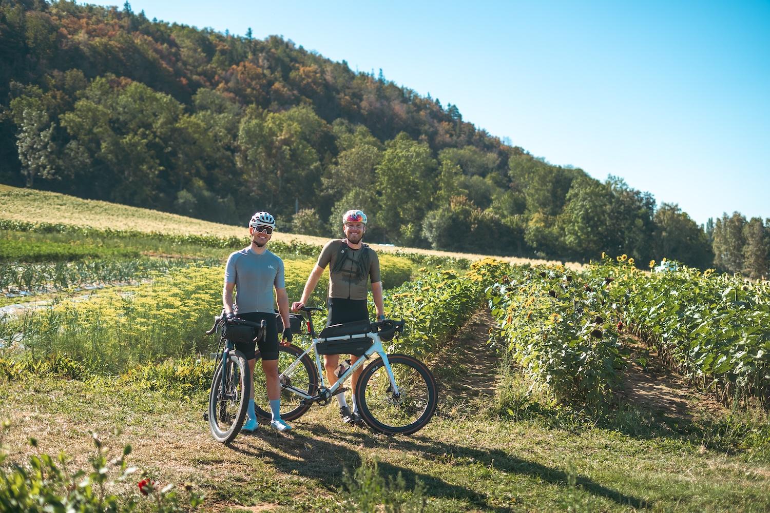 Two smiling men stand with their gravel bikes in a sunny field with a forested hill in the background.