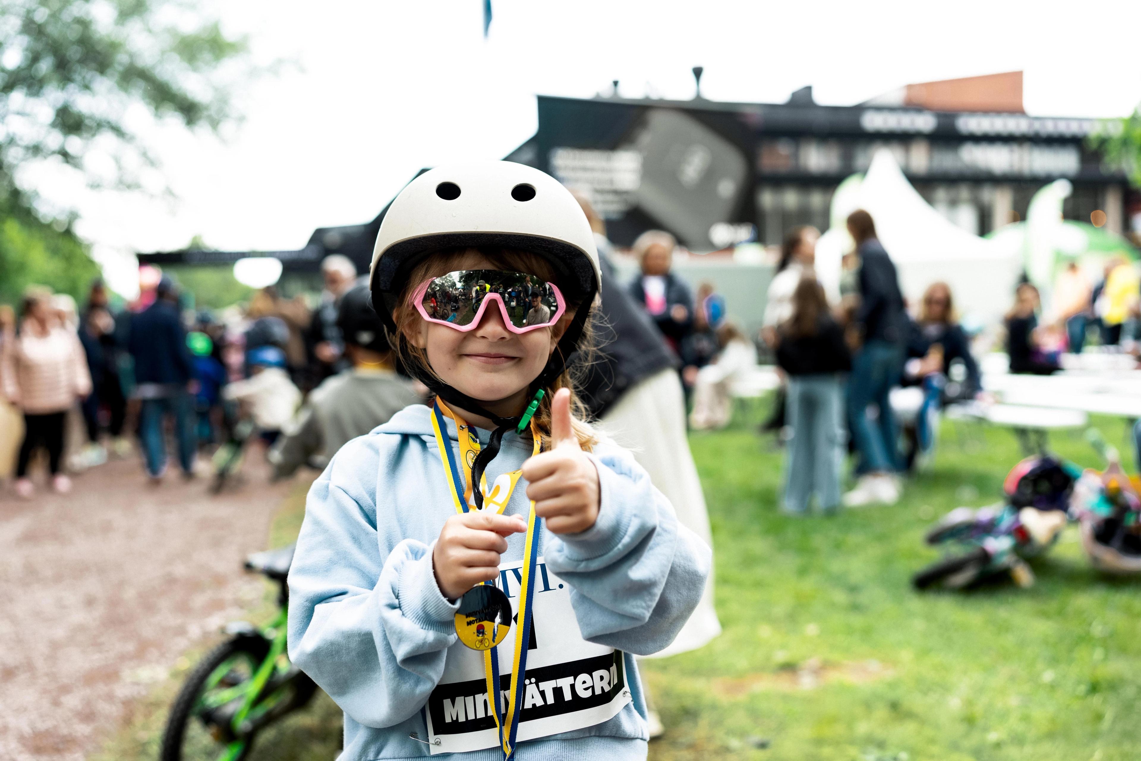 A smiling child in a bike helmet, pink sunglasses, and two medals gives a double thumbs-up at an outdoor biking event.