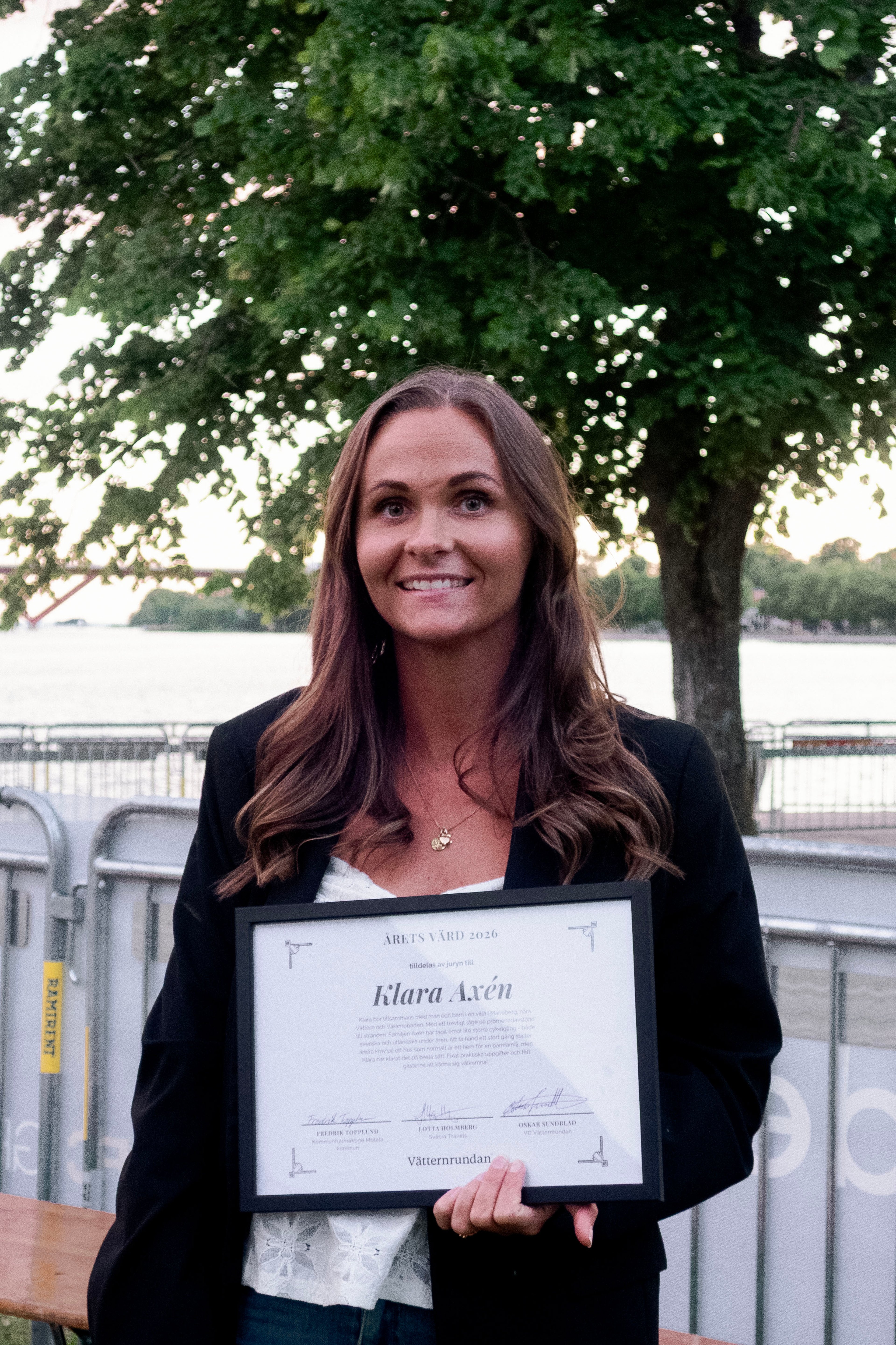 A smiling woman, Klara Axén, holds a framed certificate for "Årets Värd 2026" outdoors.