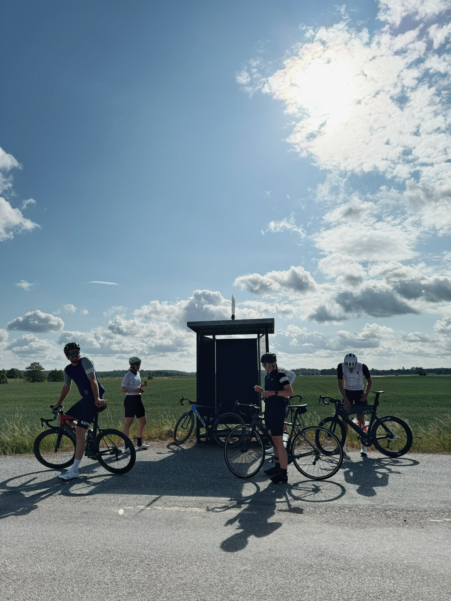 Four cyclists with road bikes stopped on a sunny rural road next to a small shelter.