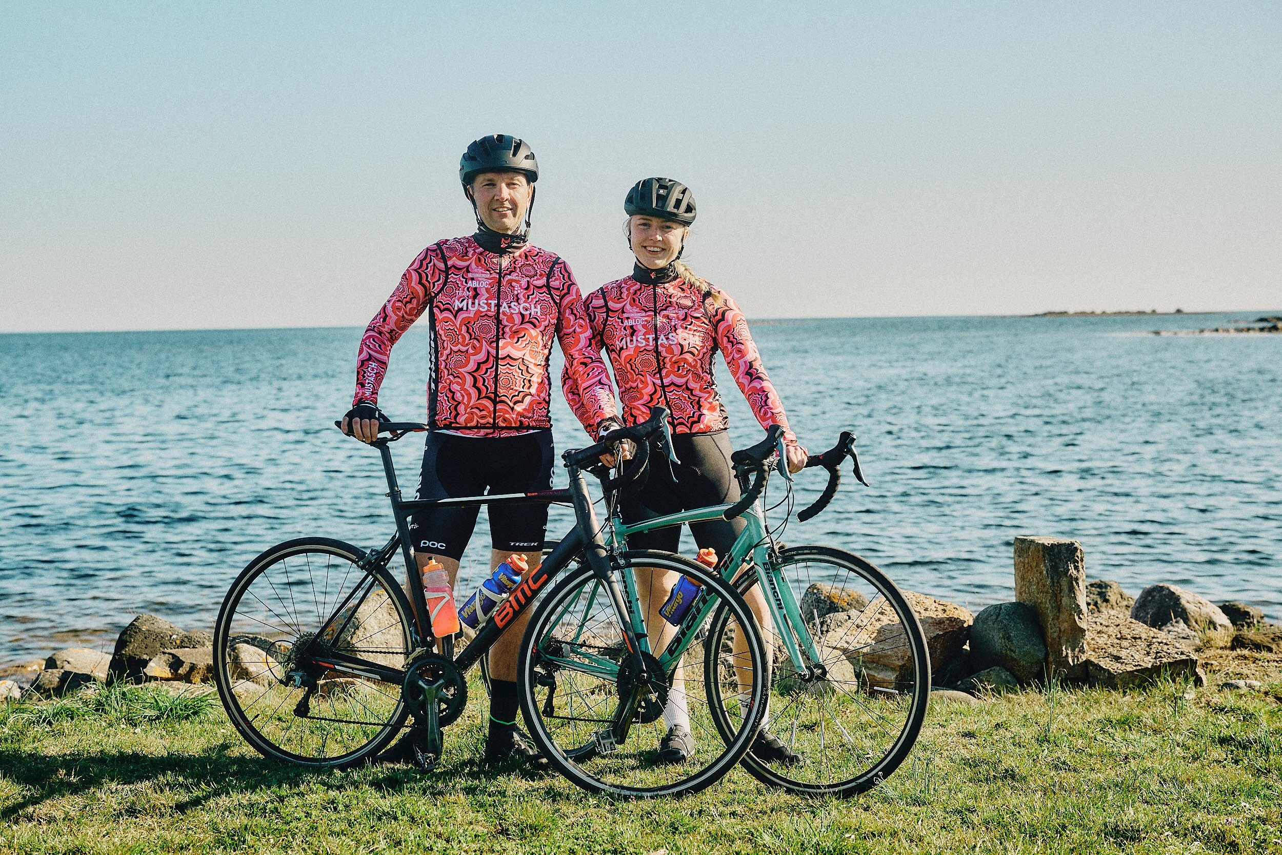 Two cyclists in matching pink patterned jerseys and helmets stand with their bikes by the ocean on a sunny day.