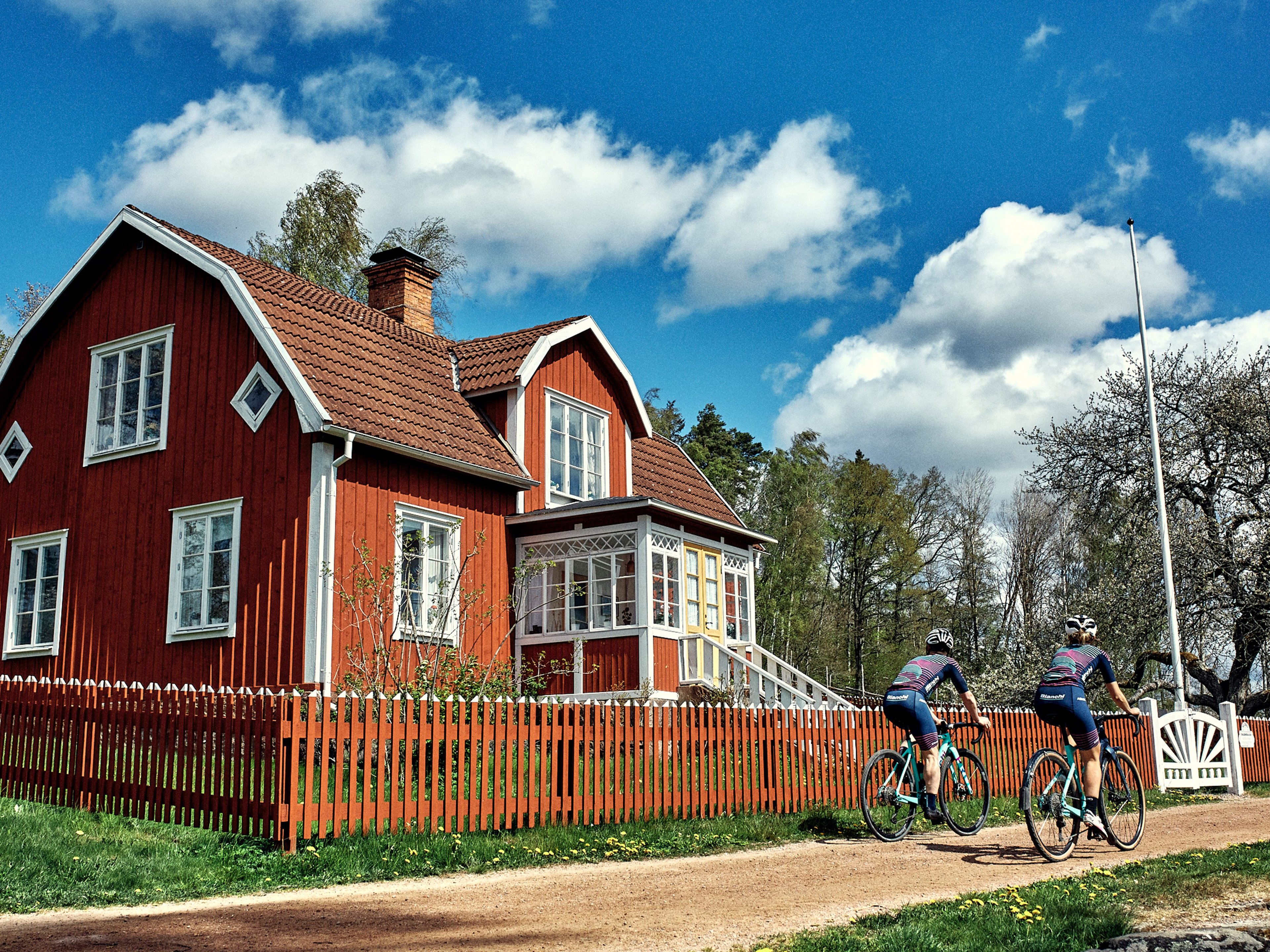 Two cyclists ride on a dirt path past a traditional red house with a picket fence.