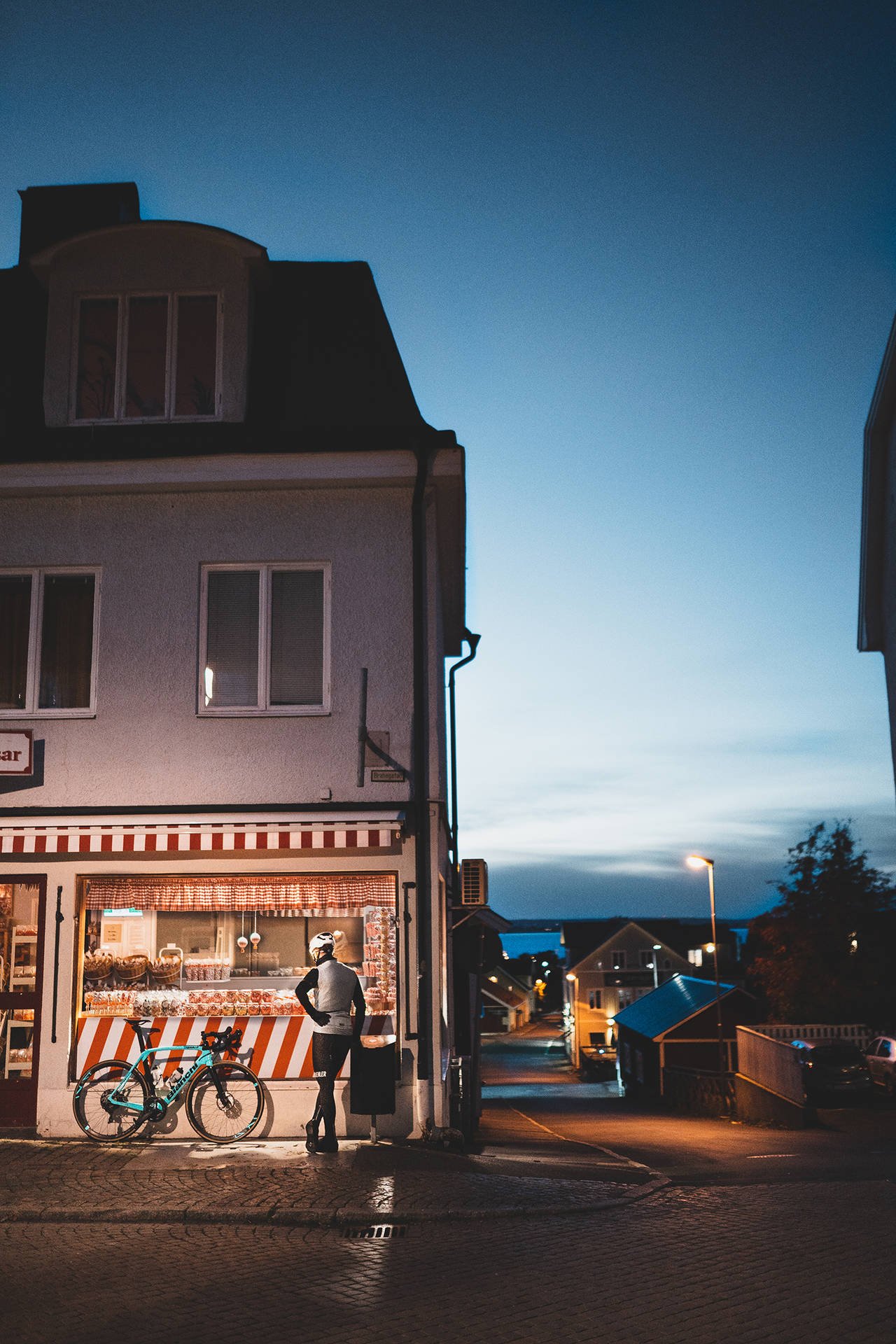 A cyclist with a turquoise bike pauses beside a brightly lit shop on a cobblestone street at dusk.