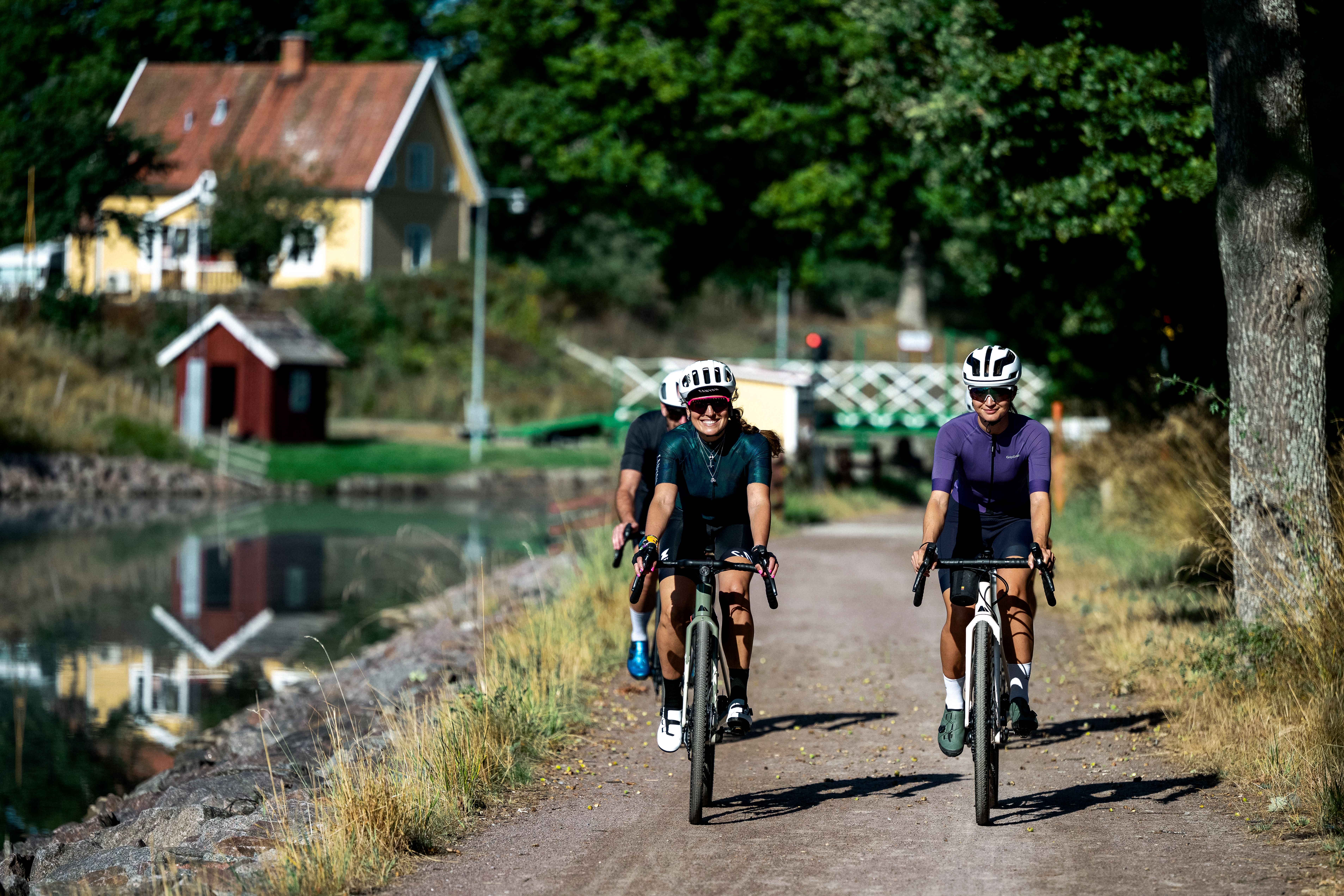 Three cyclists riding on a gravel path next to a canal with a yellow house reflecting in the water.