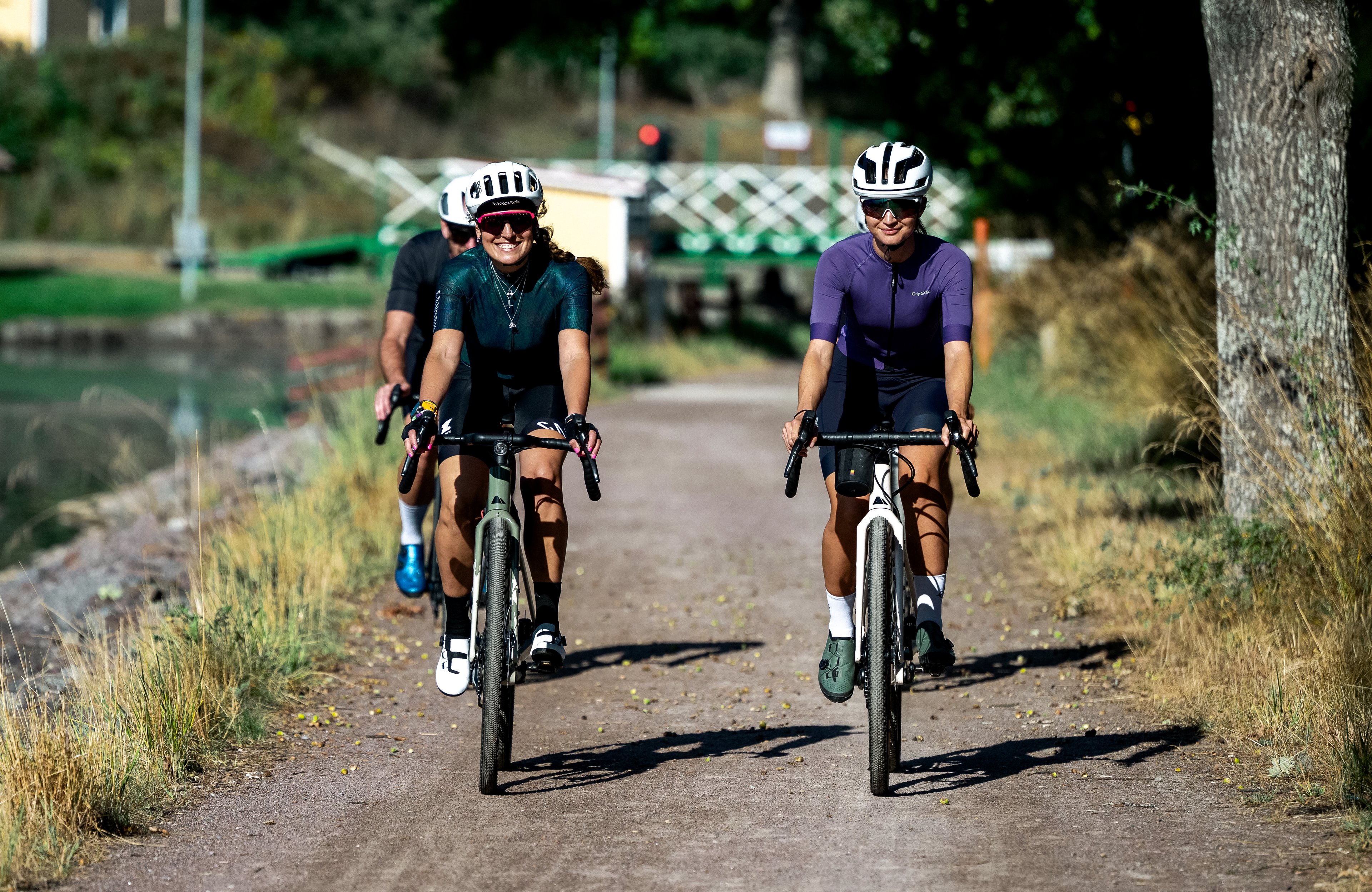 Three cyclists riding on a gravel path next to a canal with a yellow house reflecting in the water.