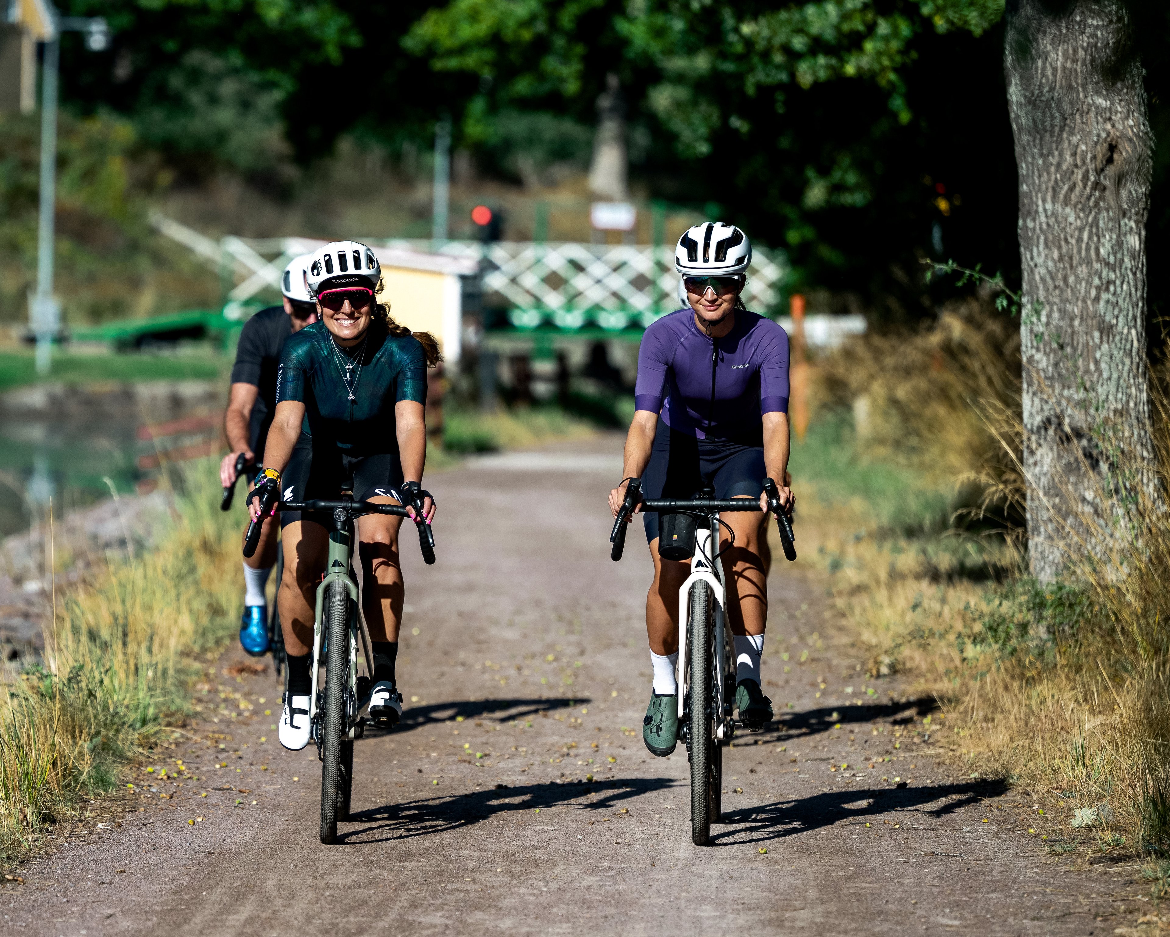 Three cyclists ride gravel bikes on a path beside a canal with a yellow house reflected in the water.