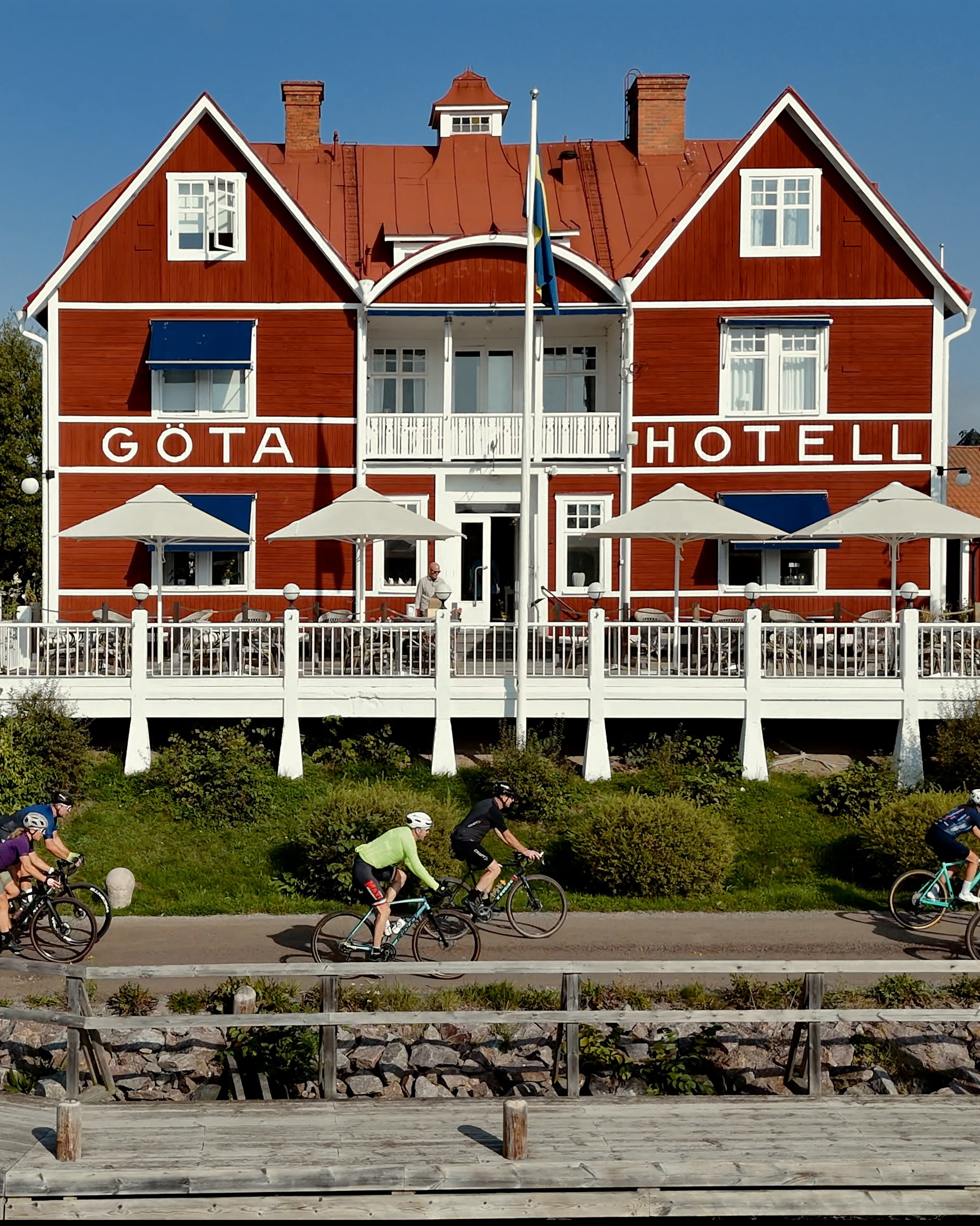 The red GÖTA HOTELL with a terrace and Swedish flag, and cyclists riding on a path below, with a wooden dock in the foreground.