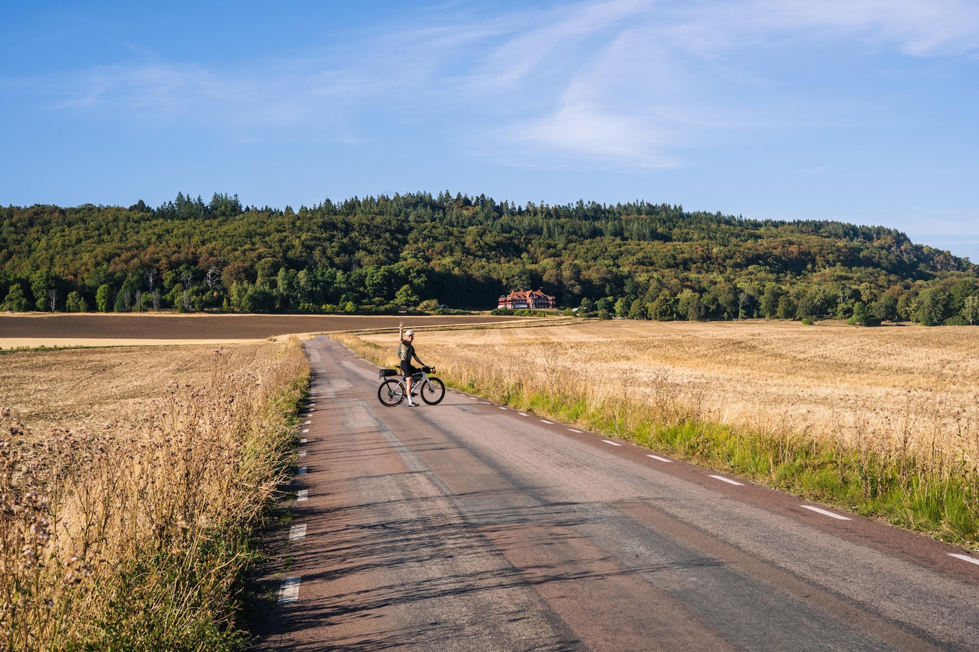 A cyclist stands with their bike on a rural road, golden fields stretching to a forested hill with a distant house.
