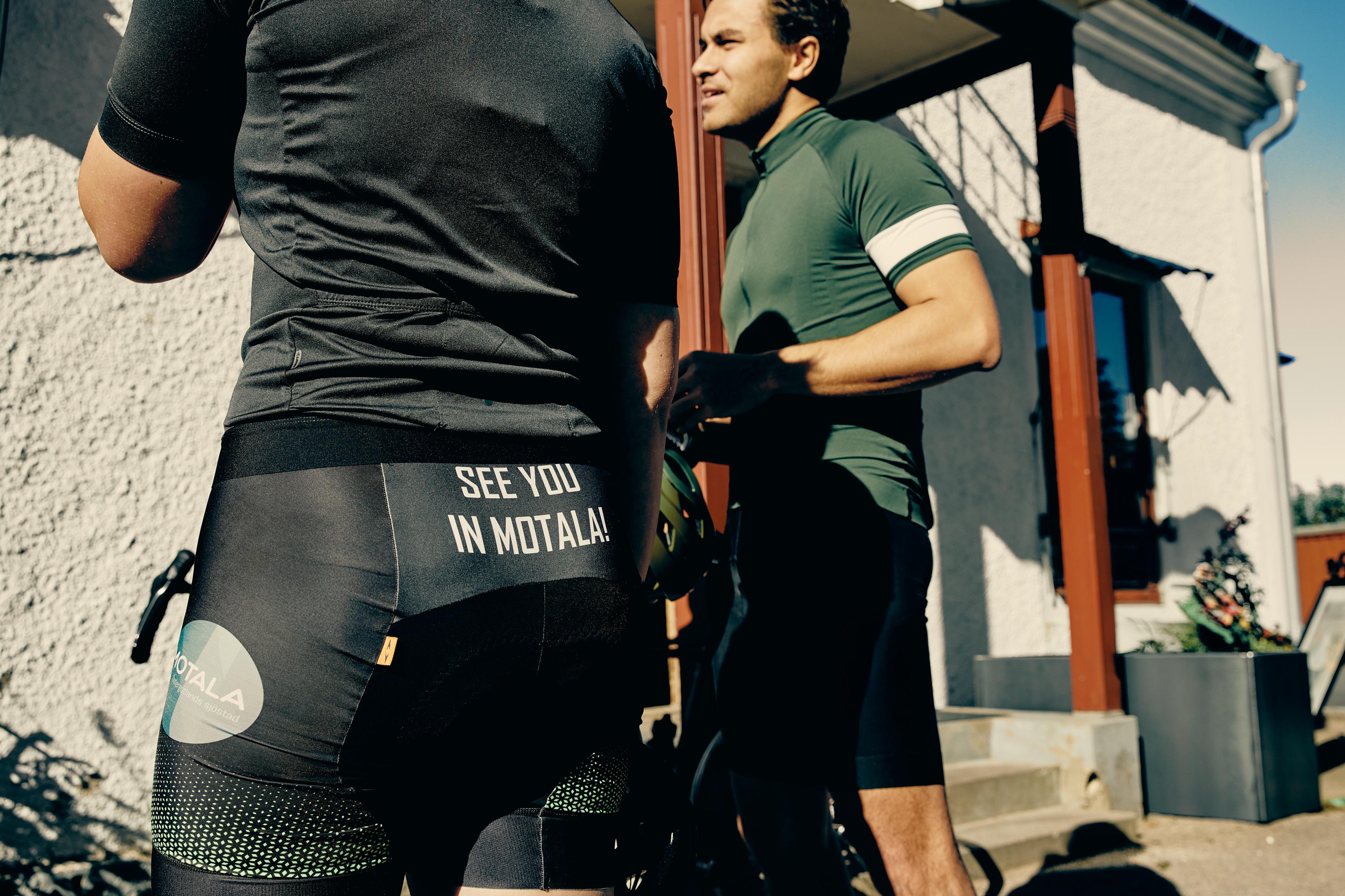 Two cyclists outdoors; one seen from behind wearing shorts with "SEE YOU IN MOTALA!", the other in a green jersey.