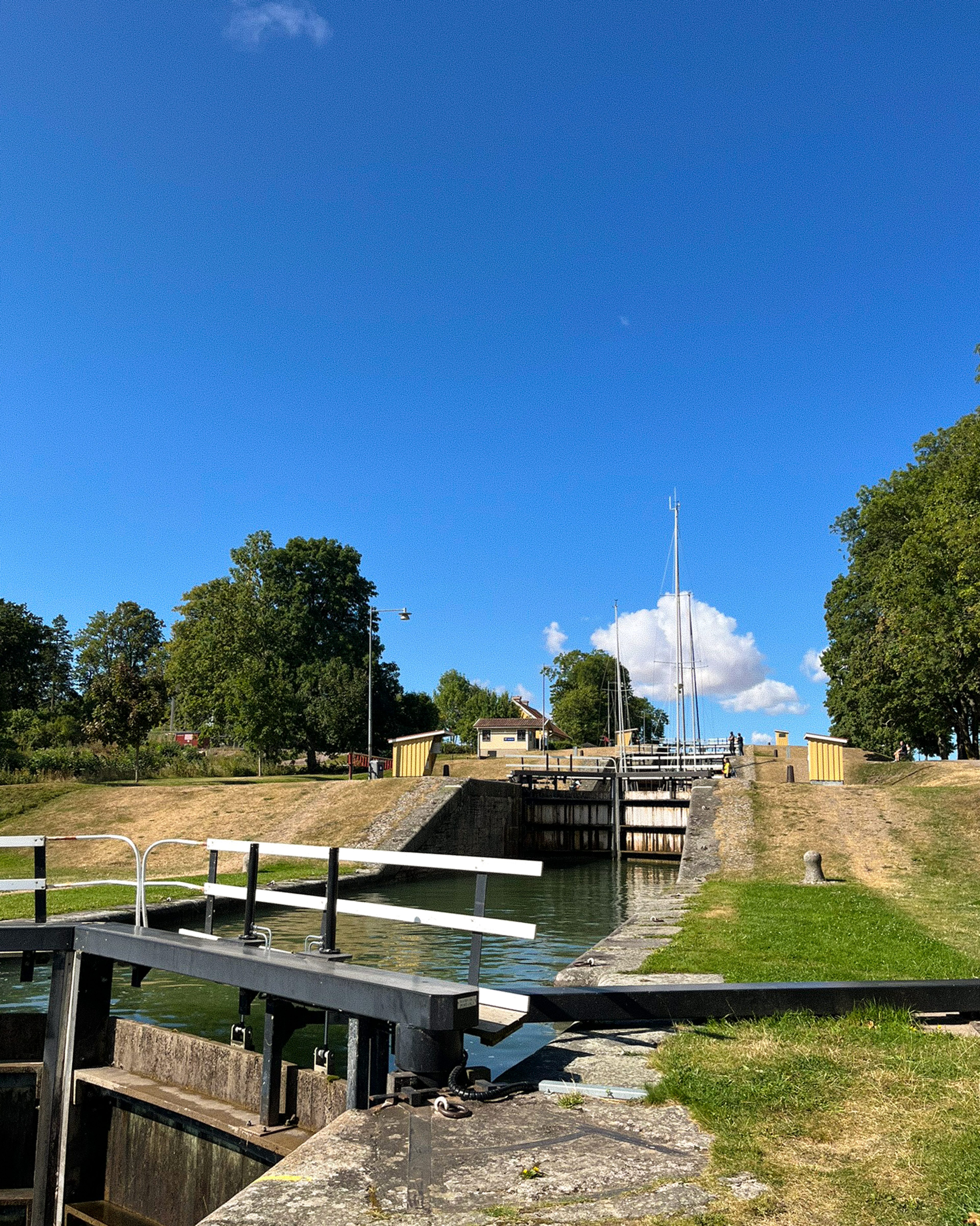 A sunny view of a canal lock system with open gates, green water, grassy banks, trees, and a sailboat mast in the distance under a bright blue sky.