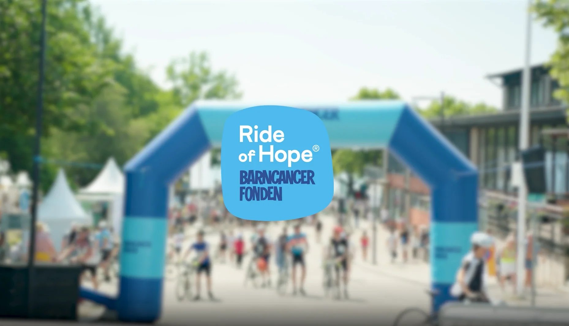 A blurred image of a cycling event with an inflatable arch, overlaid with a blue oval logo reading "Ride of Hope® BARNCANCER FONDEN".