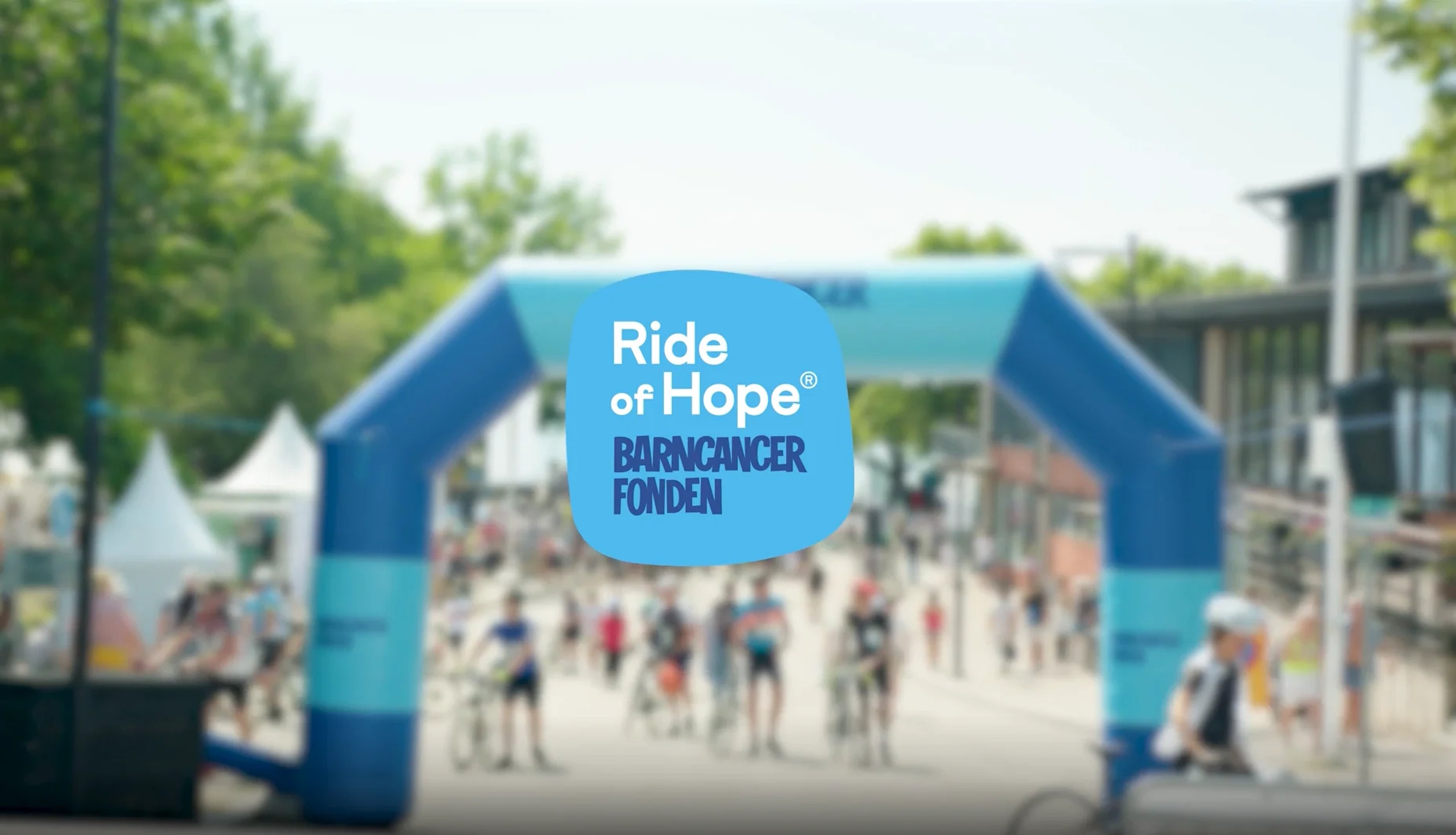 A blurred image of a cycling event with an inflatable arch, overlaid with a blue oval logo reading "Ride of Hope® BARNCANCER FONDEN".