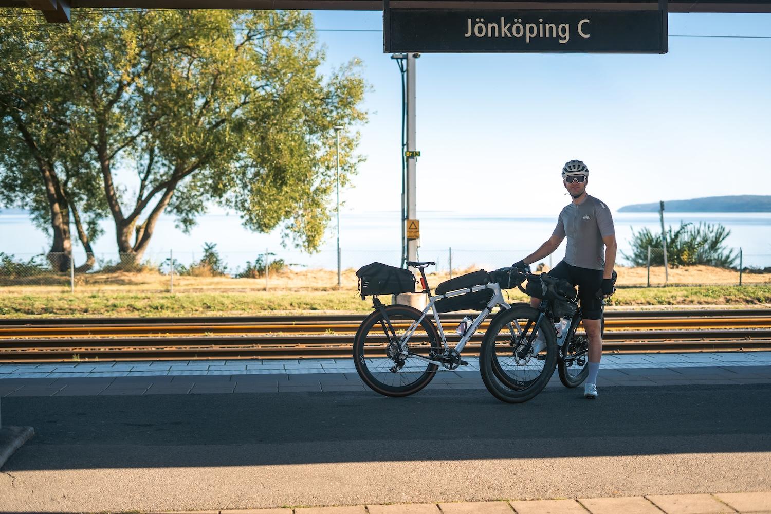 A man and two touring bikes on a sunny train platform, with a "Jönköping C" sign and a lake view.