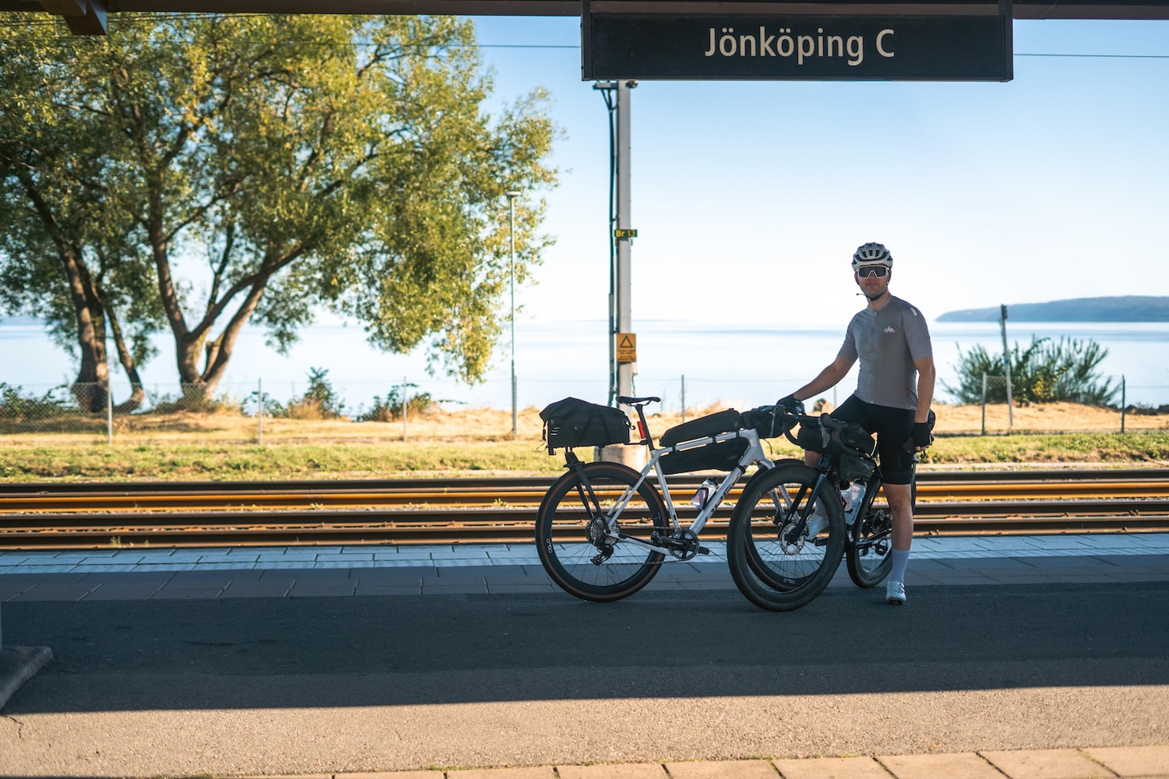 A man and two touring bikes on a sunny train platform, with a "Jönköping C" sign and a lake view.