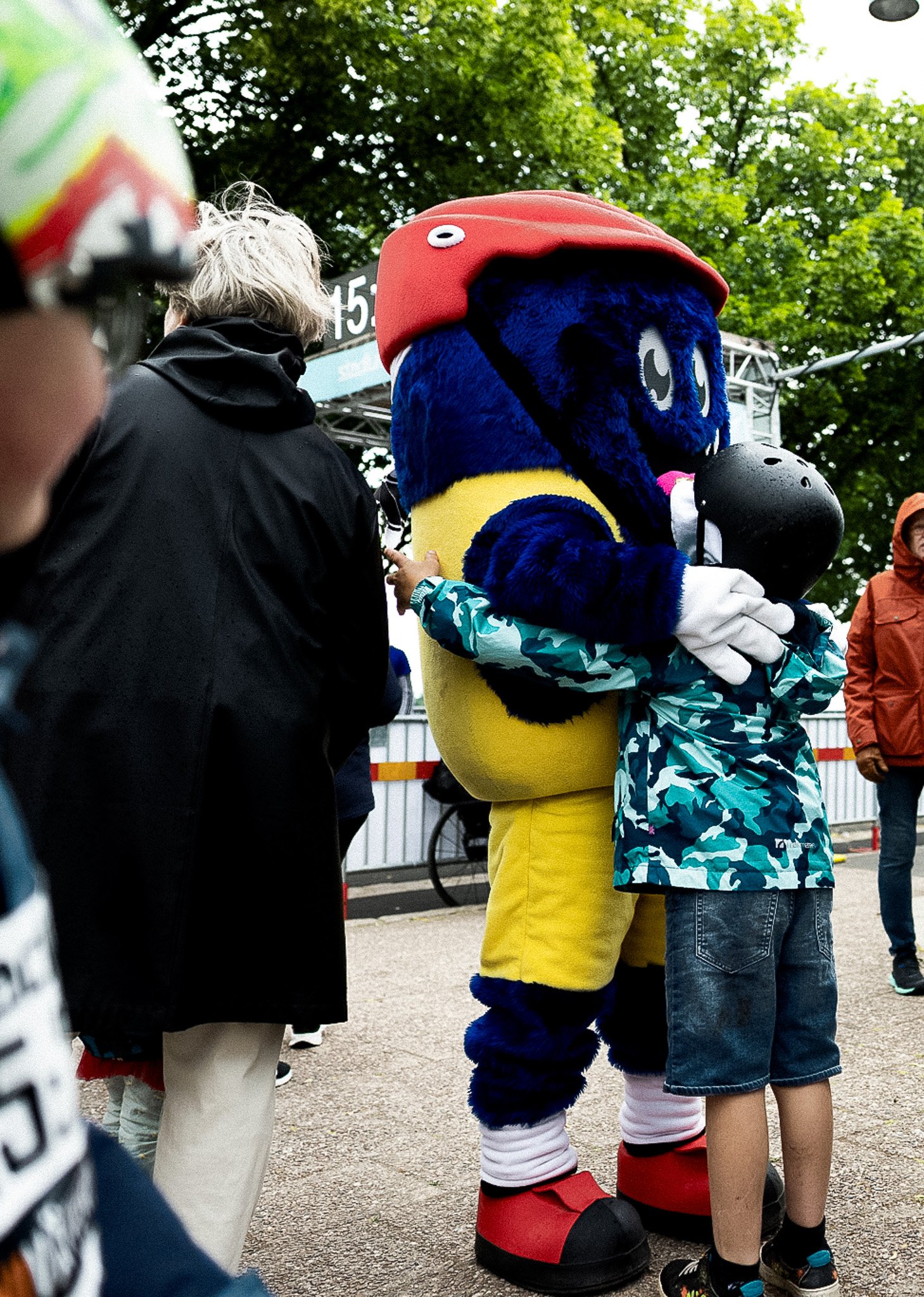 A blue and yellow mascot with a red helmet hugs a child wearing a camouflage jacket and black helmet.