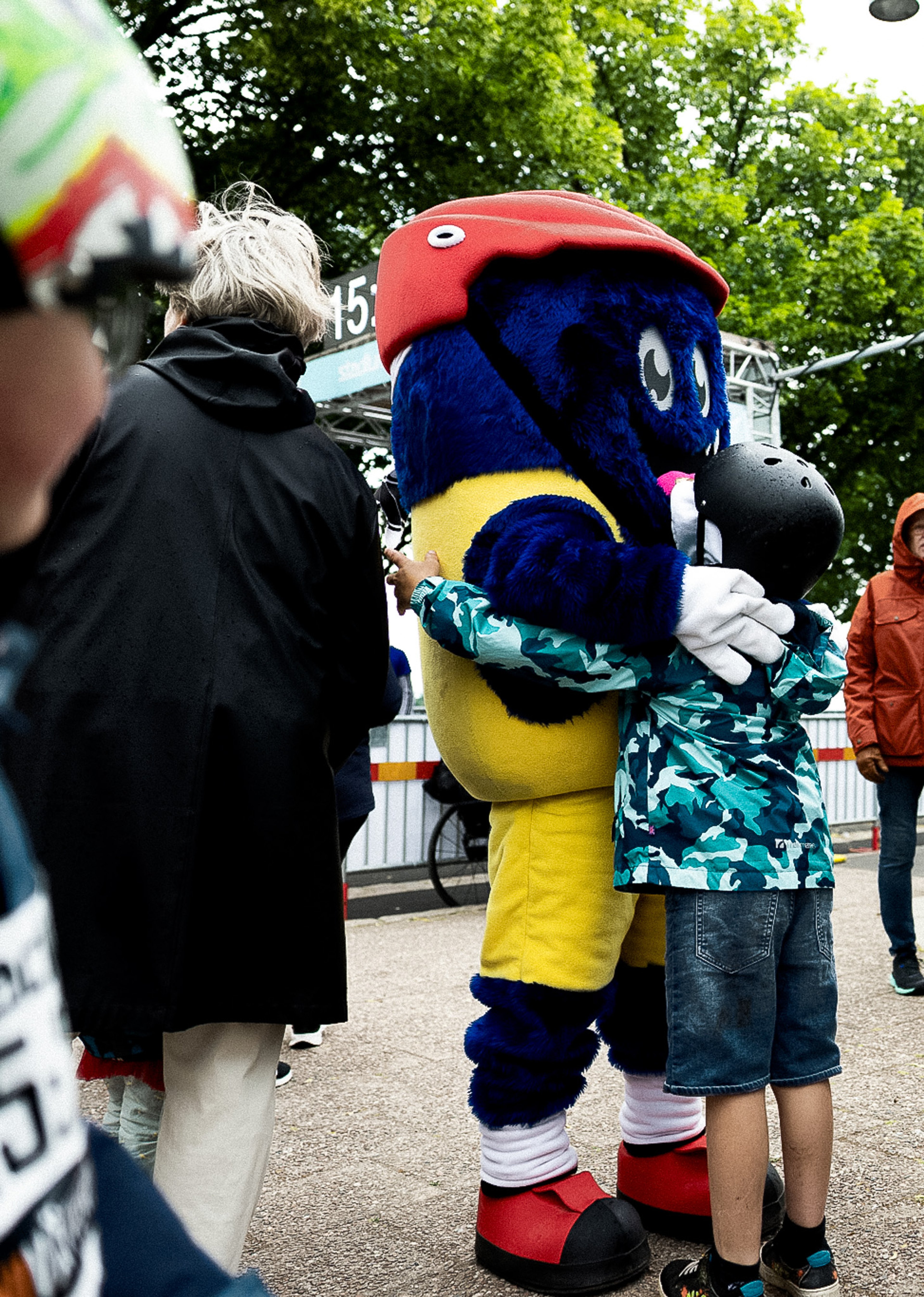 A blue and yellow mascot with a red helmet hugs a child wearing a camouflage jacket and black helmet.