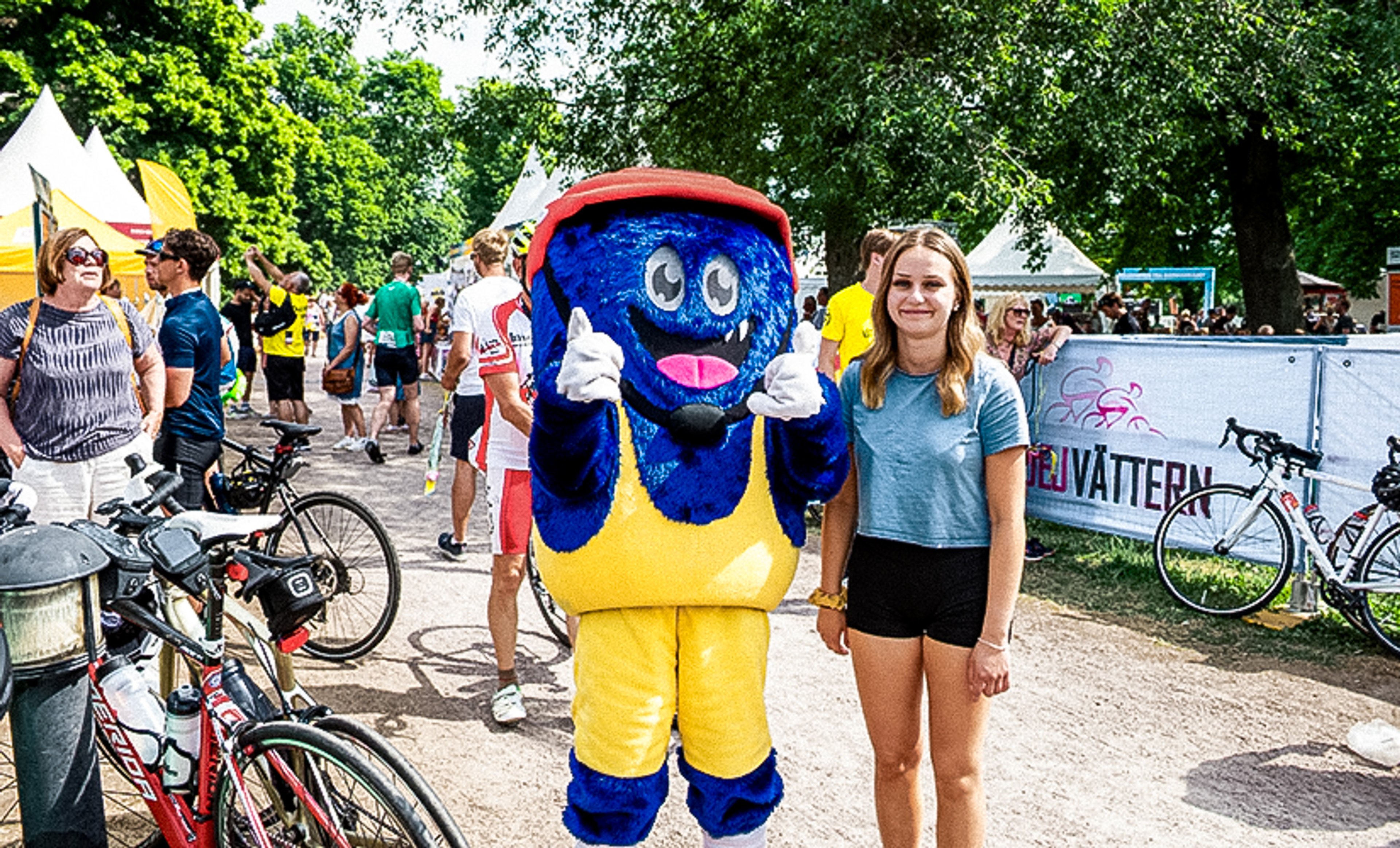 A blue furry mascot in yellow overalls and a red cap gives two thumbs up next to a young woman at a cycling event.