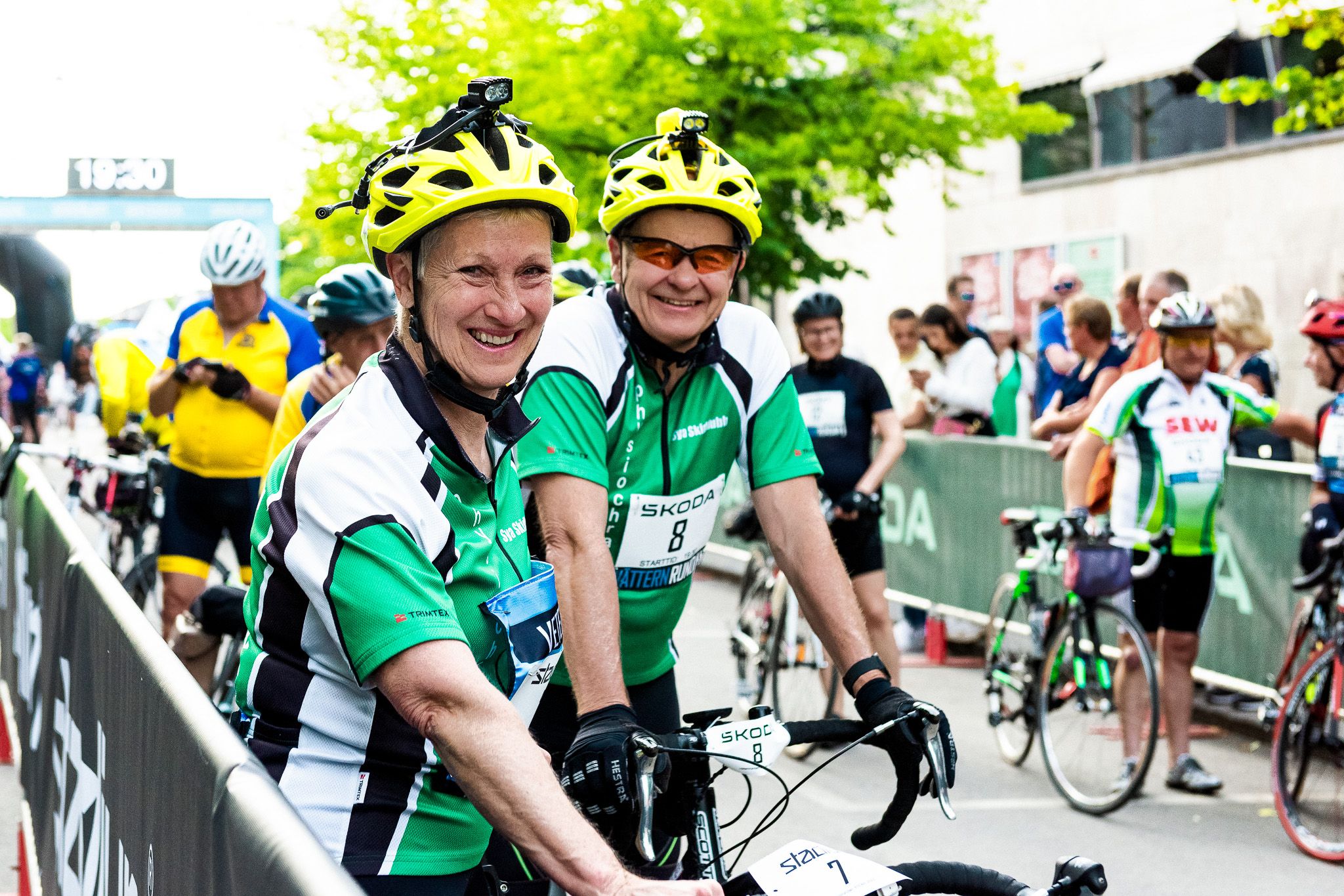 Two smiling cyclists in yellow helmets and green jerseys stand with their bikes at a race event.