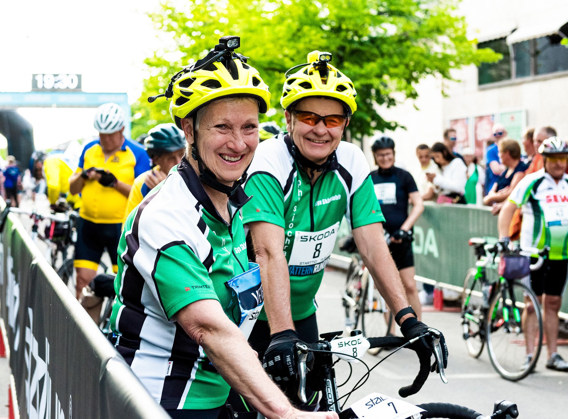 Two smiling cyclists in yellow helmets and green jerseys stand with their bikes at a race event.