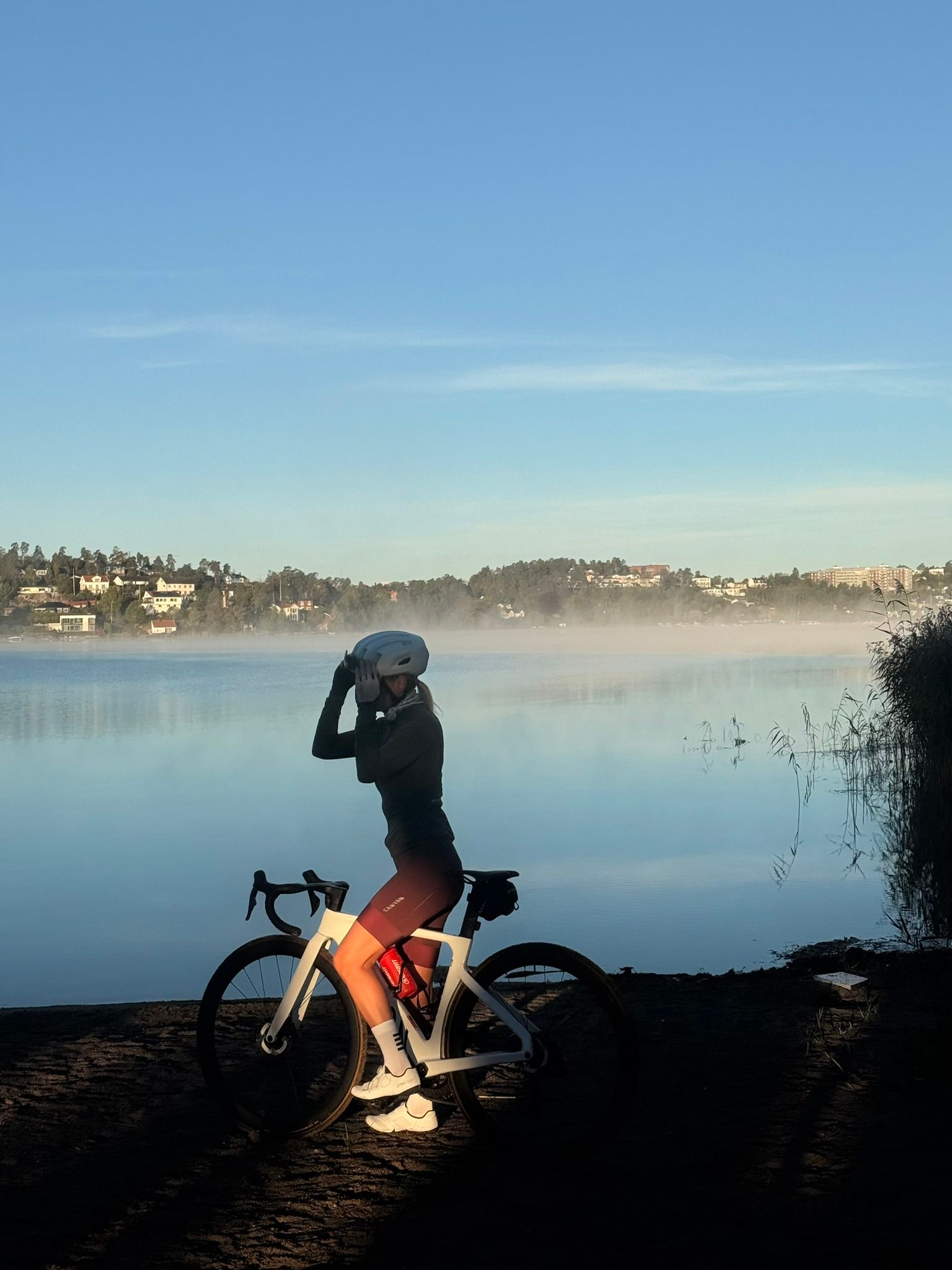 A cyclist stands with a white bike on the dark shore of a misty lake under a clear blue sky.