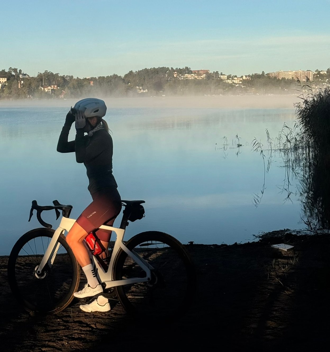 A cyclist stands with a white bike on the dark shore of a misty lake under a clear blue sky.