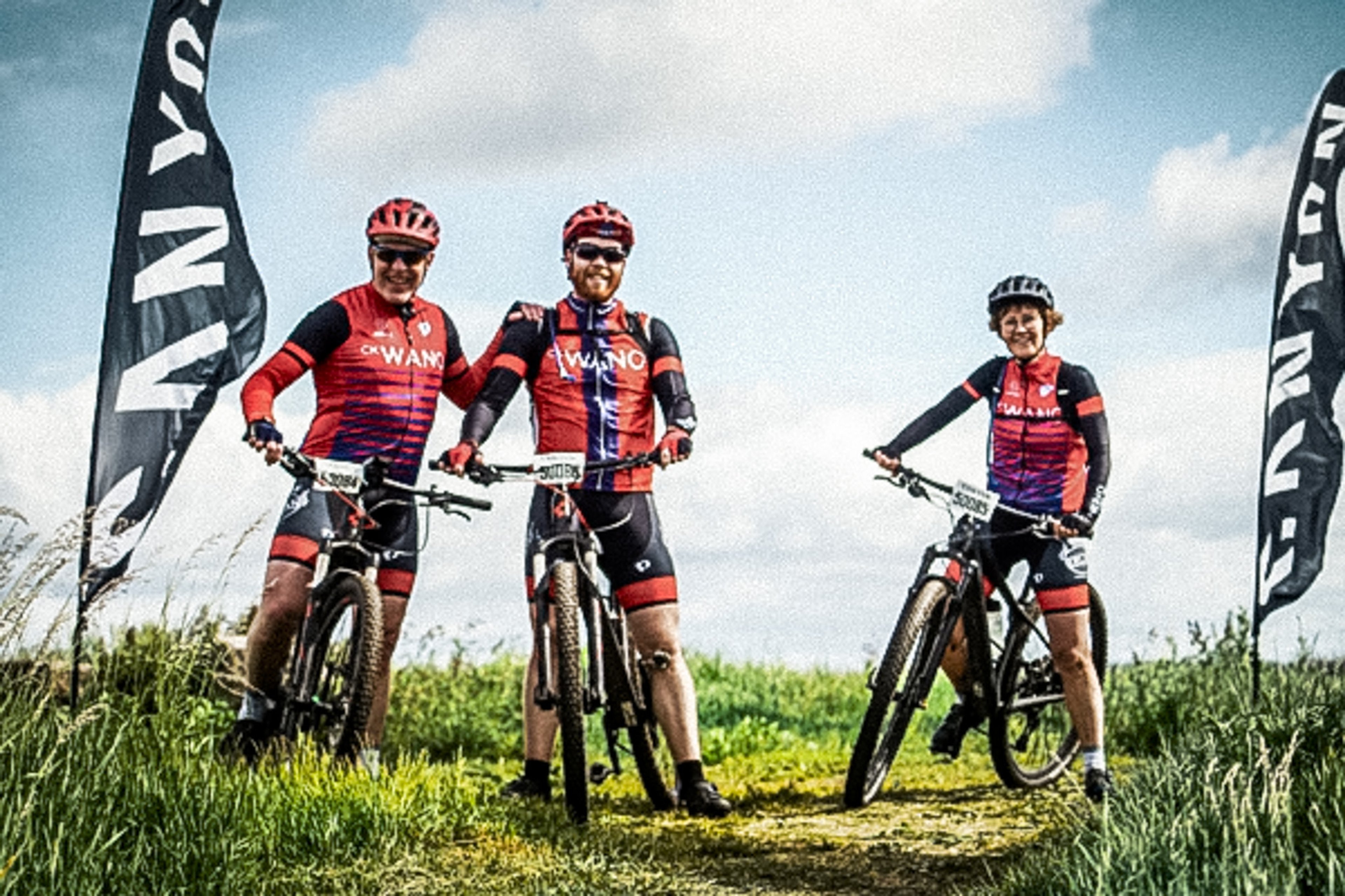 Three smiling cyclists in red and black gear stand with their mountain bikes on a grassy path between event flags.