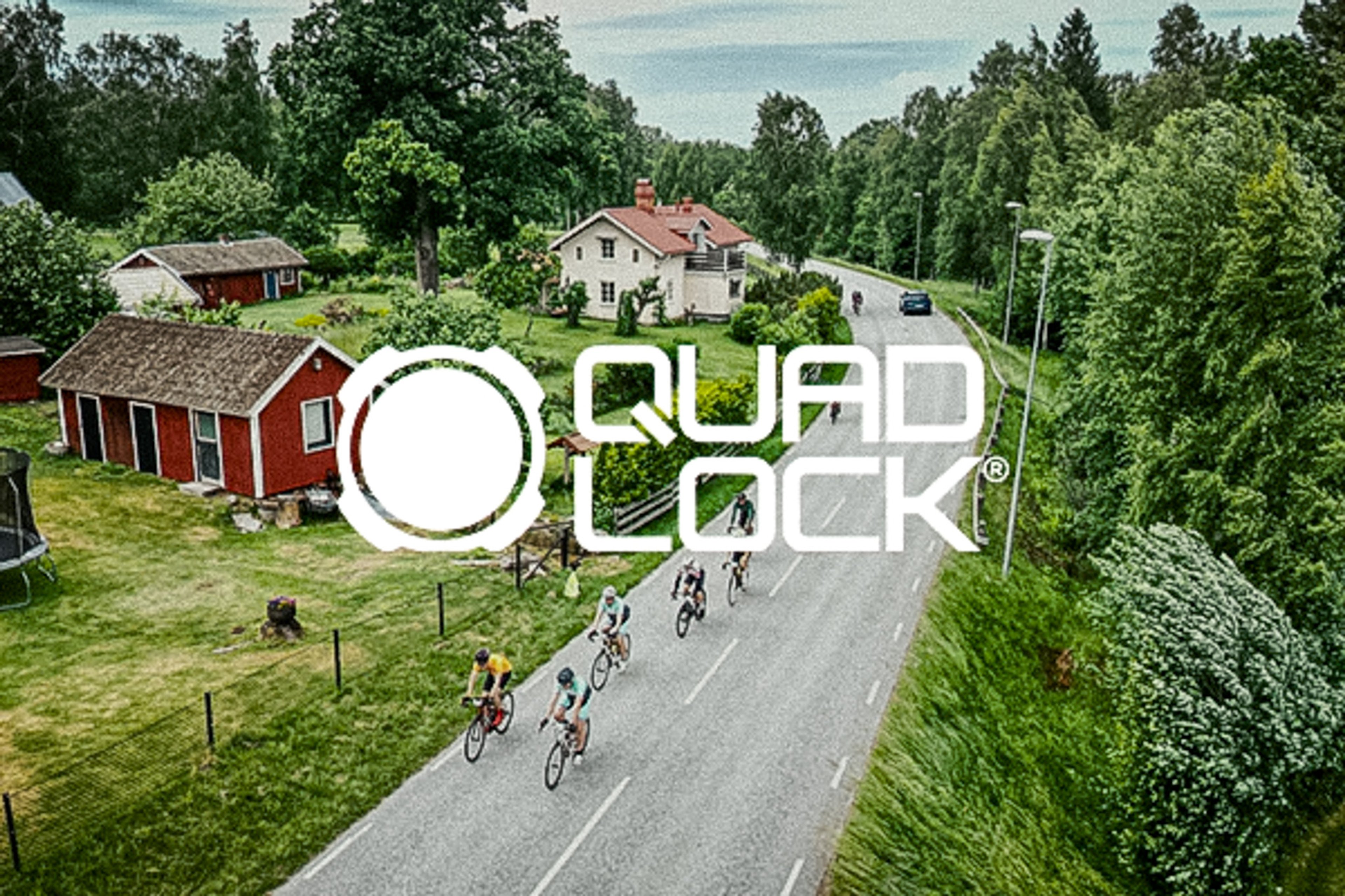 A group of cyclists rides on a country road with houses and trees, overlaid with the Quad Lock logo.