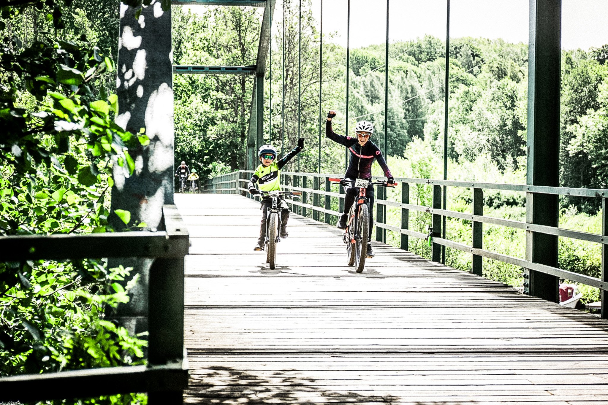 Two cyclists celebrate with raised arms on a wooden bridge surrounded by trees.