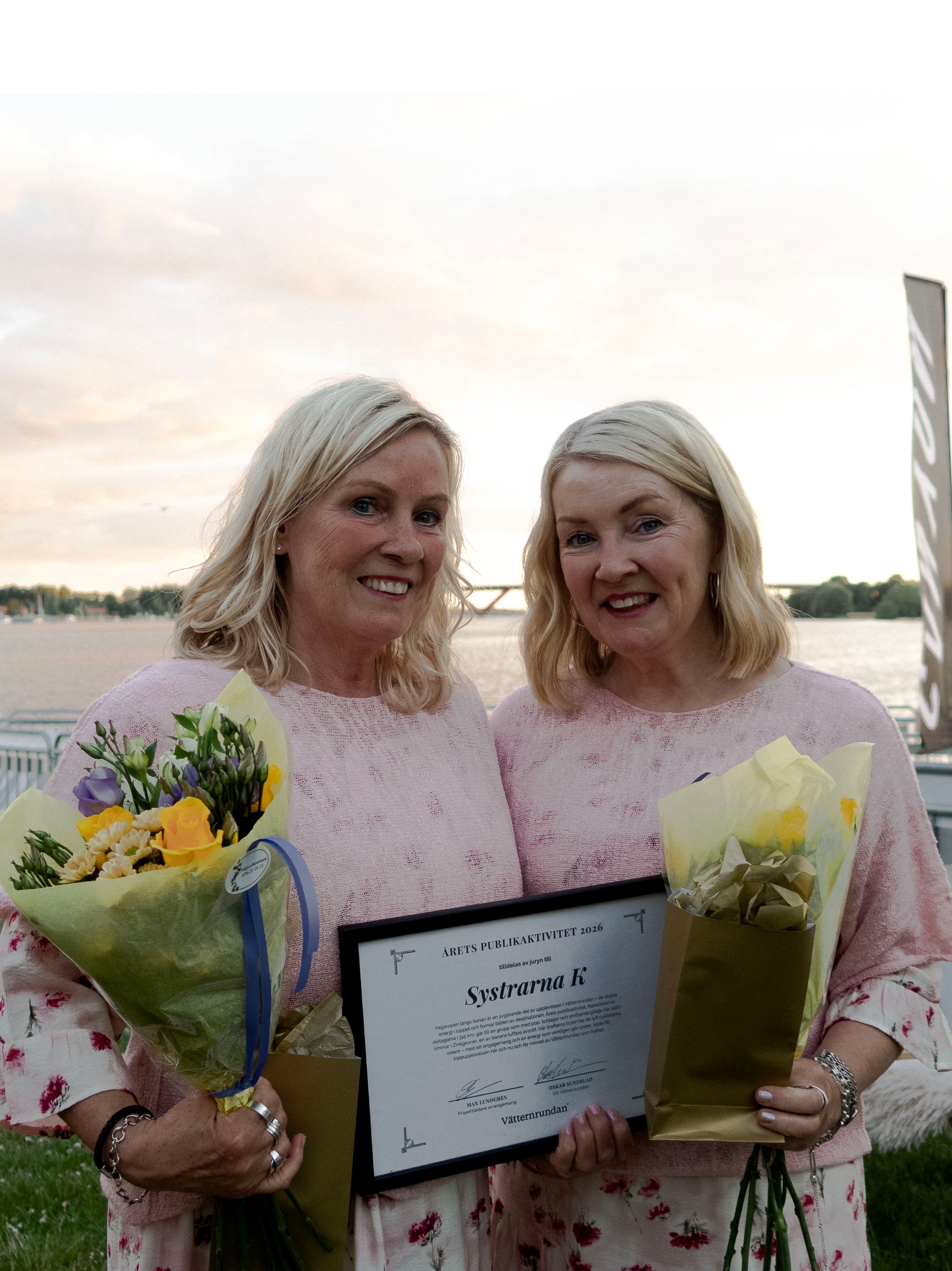Two smiling blonde women hold flower bouquets and an award certificate for "Systrarna K" by a body of water.