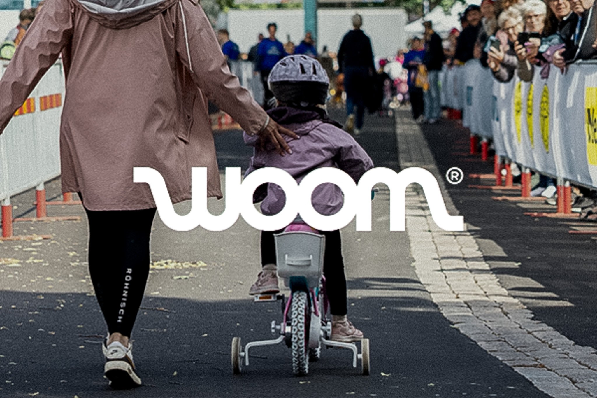 An adult guides a child on a bicycle with training wheels at an outdoor event with spectators, overlaid with the white "woom" logo.