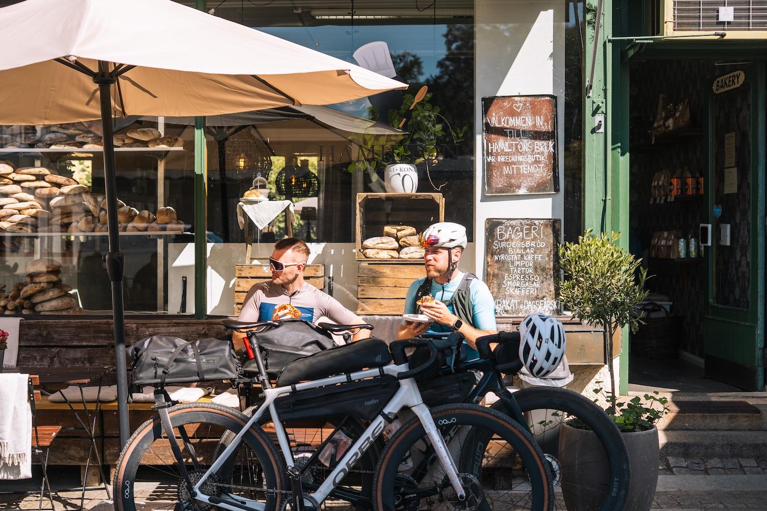 Two cyclists take a break, eating at an outdoor table in front of a bakery with their bikes parked nearby.