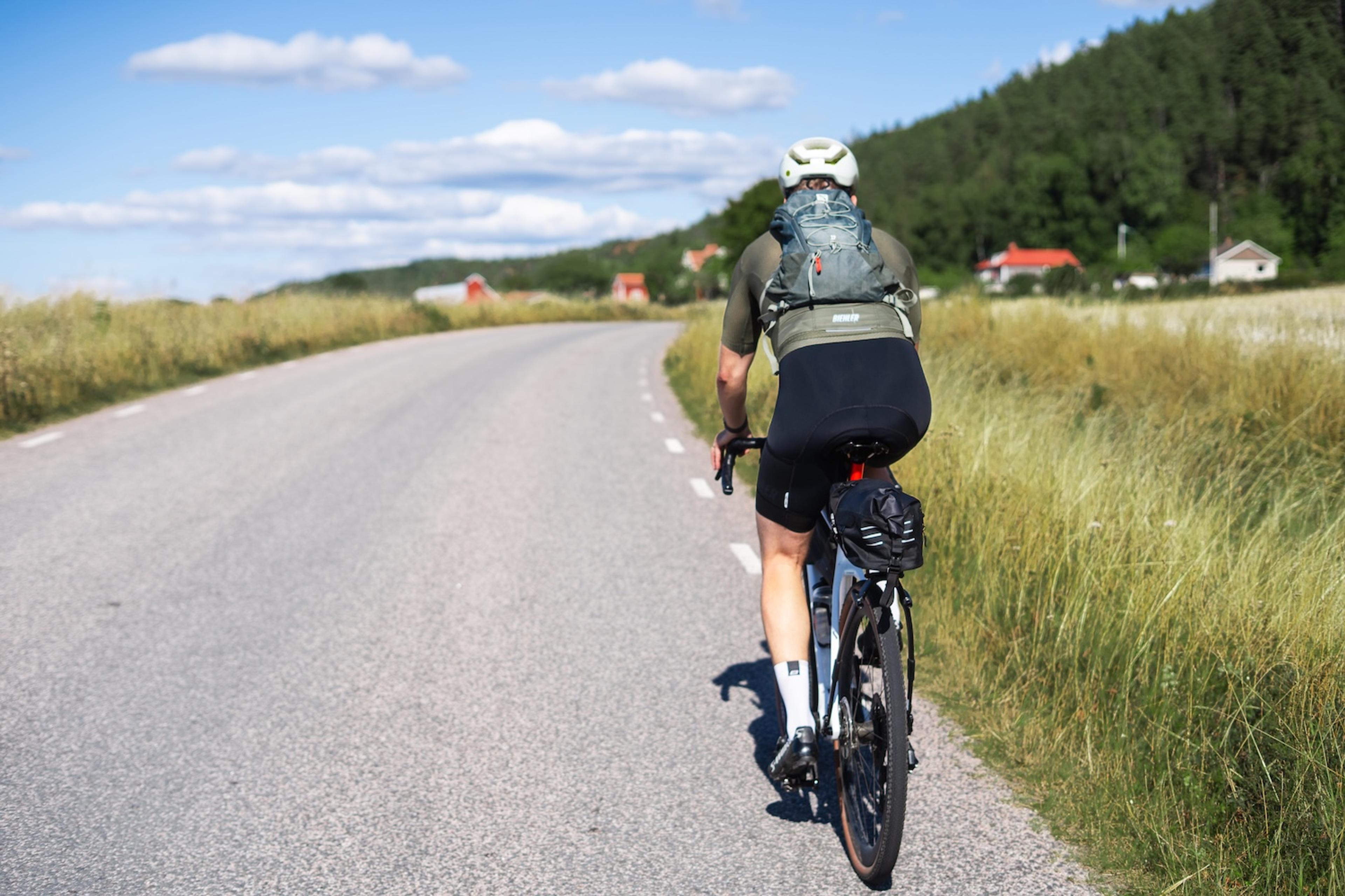 A person cycles away on a rural road, bordered by tall grass and distant houses, under a sunny sky.