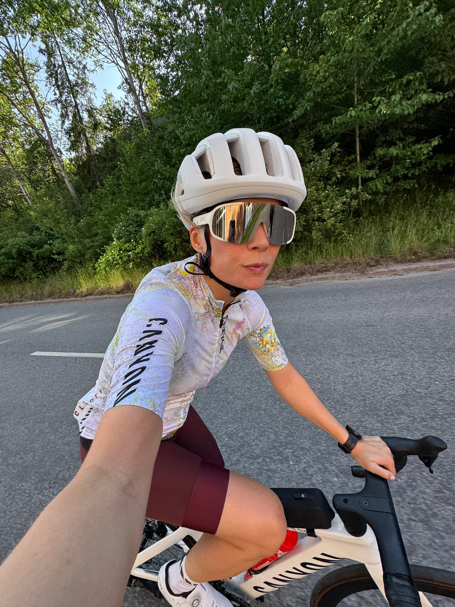 A woman in a white helmet, reflective sunglasses, and cycling kit rides a road bike on a tree-lined road.