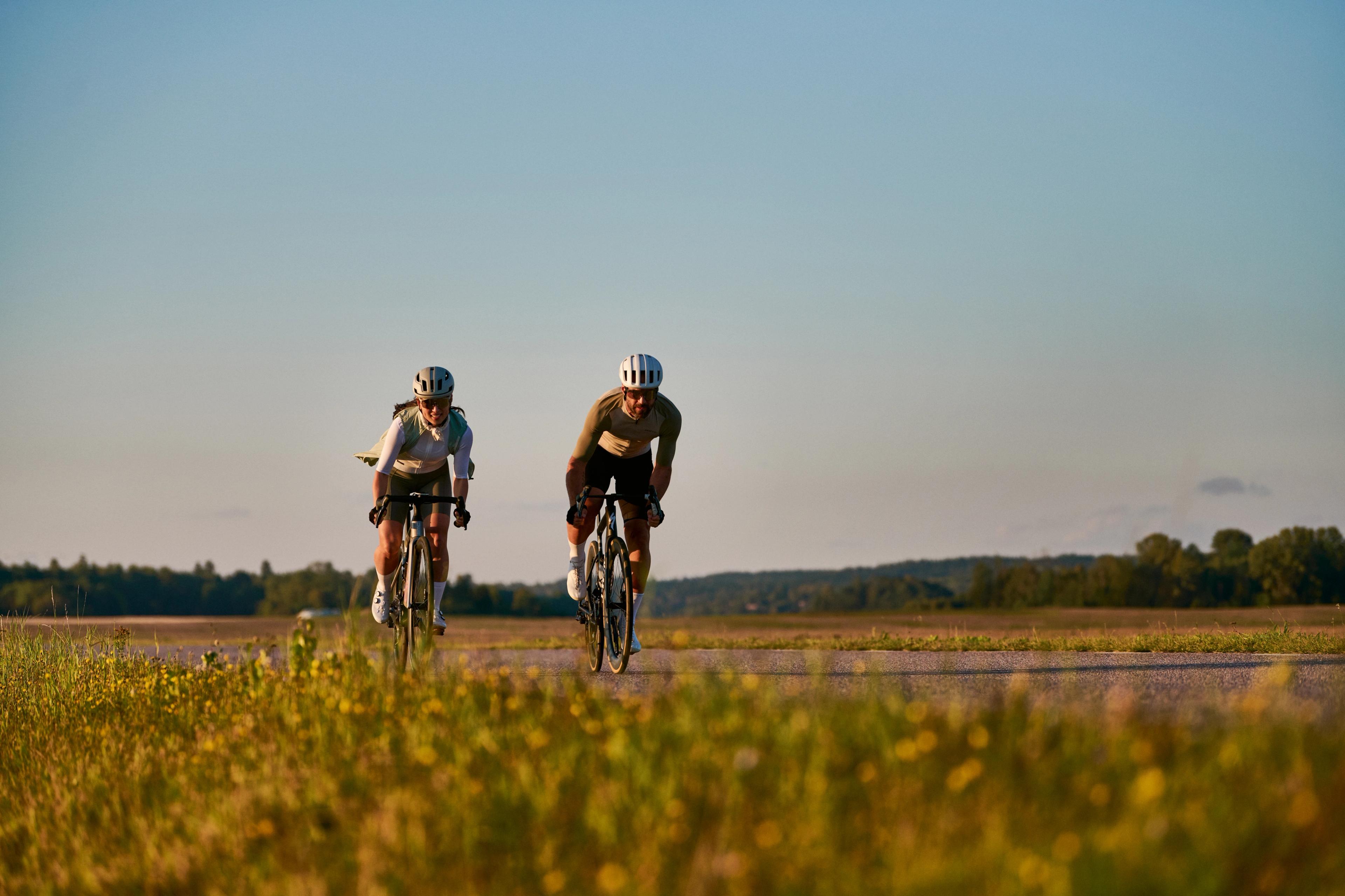 Three cyclists ride across a dry field with a dense forest under a cloudy sky.