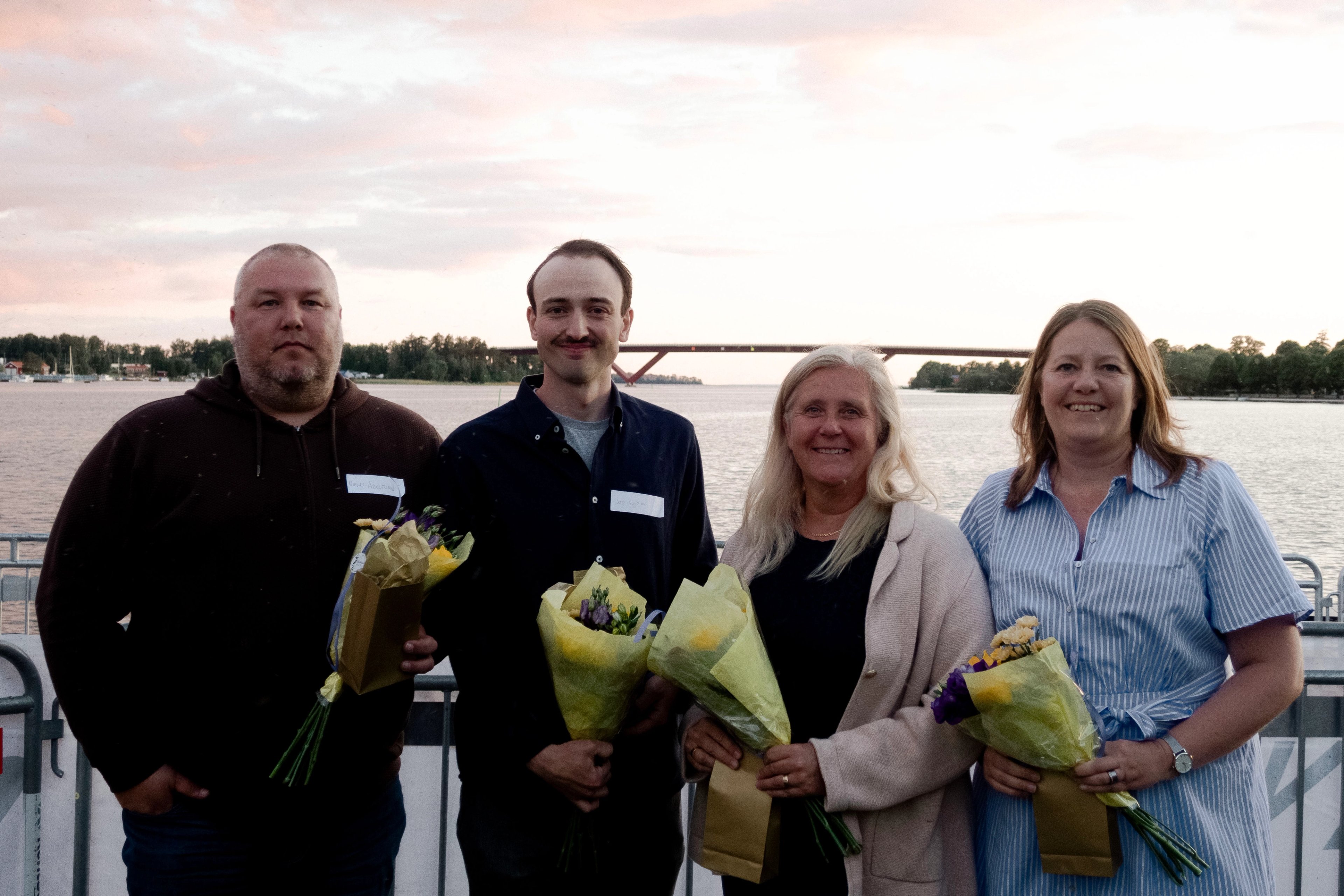 Four people, two men and two women, holding bouquets of flowers by a body of water with a bridge in the background.