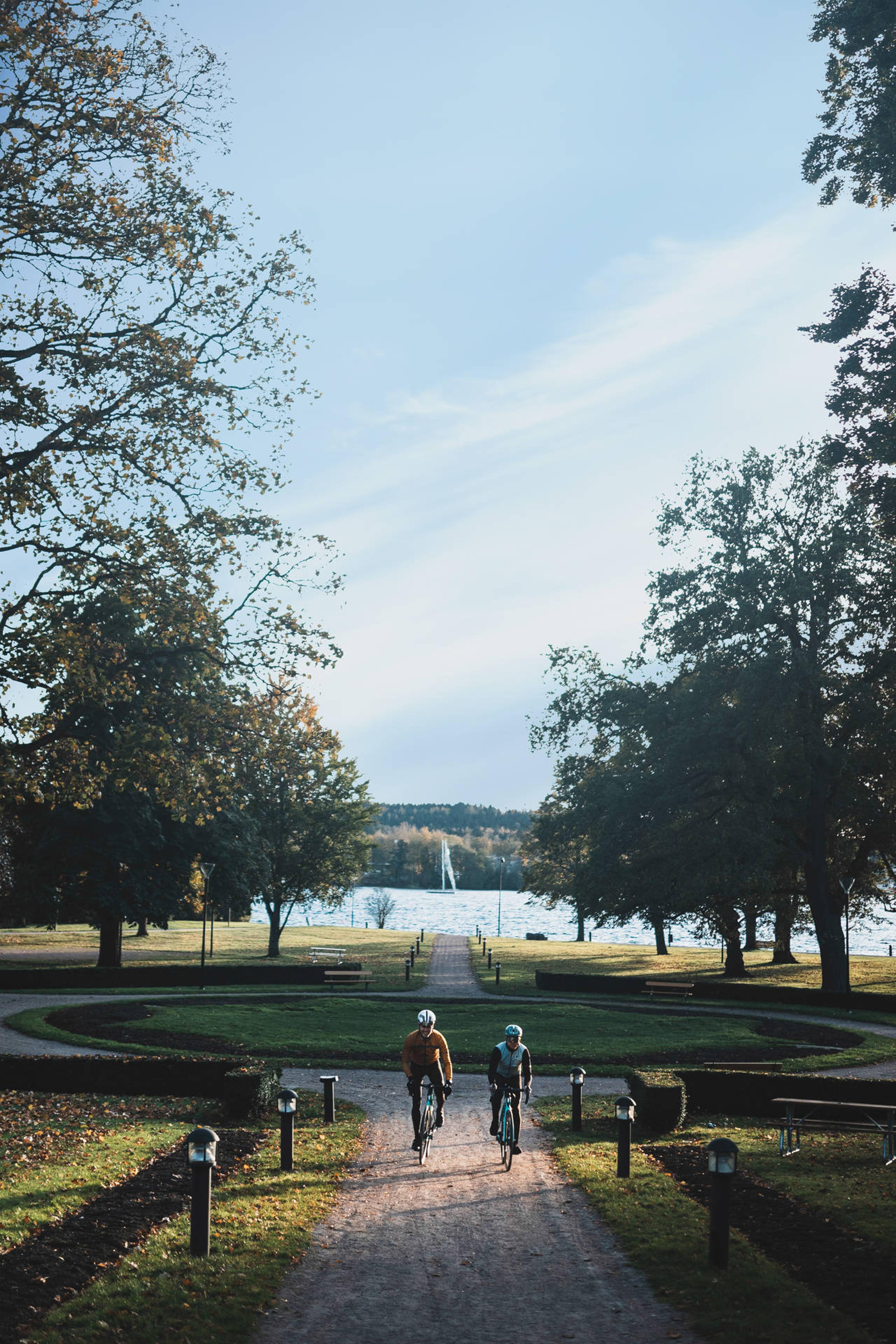 Two cyclists ride on a scenic park path leading to a lake with a sailboat.