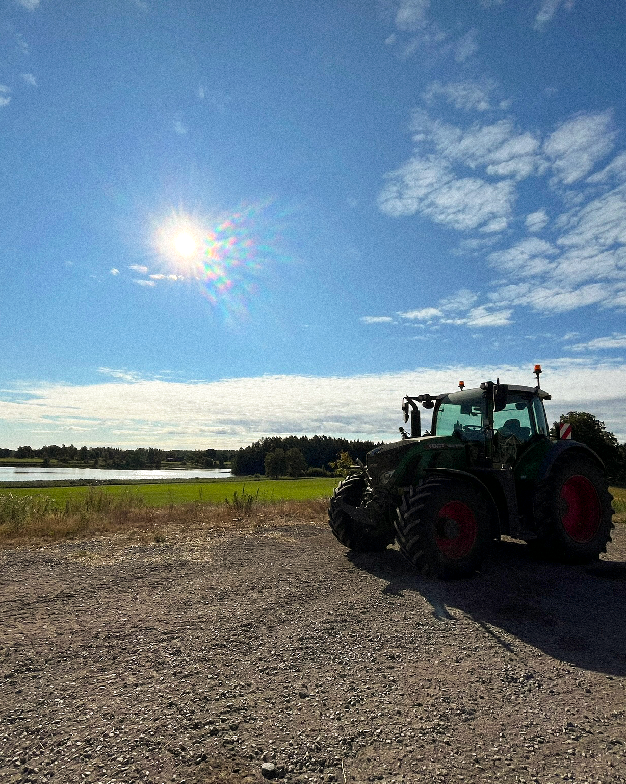 A green tractor on a gravel path overlooking a lake and fields under a bright, sunny blue sky.