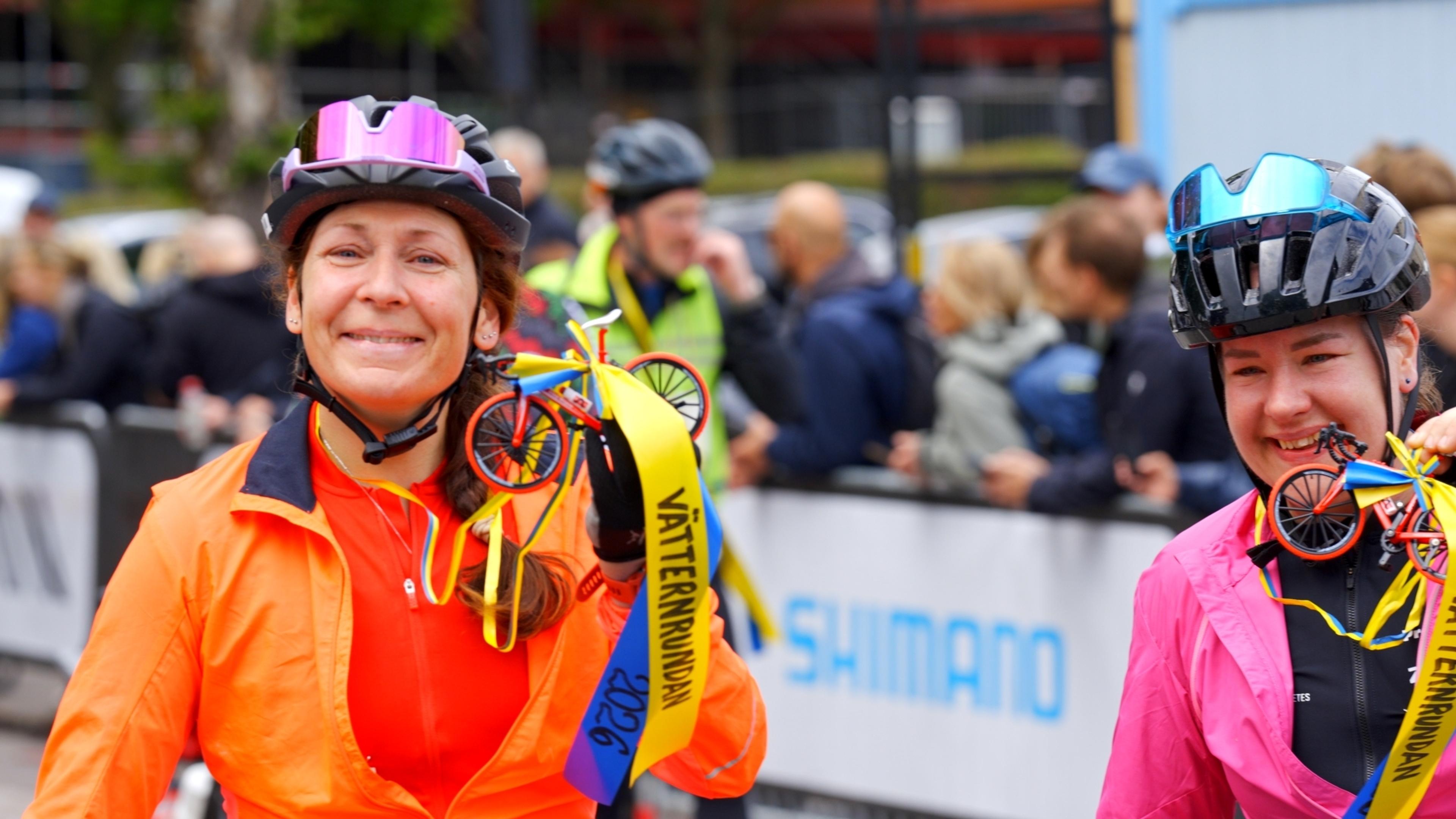 Two smiling women in cycling helmets holding bicycle-shaped medals with Vätternrundan ribbons.