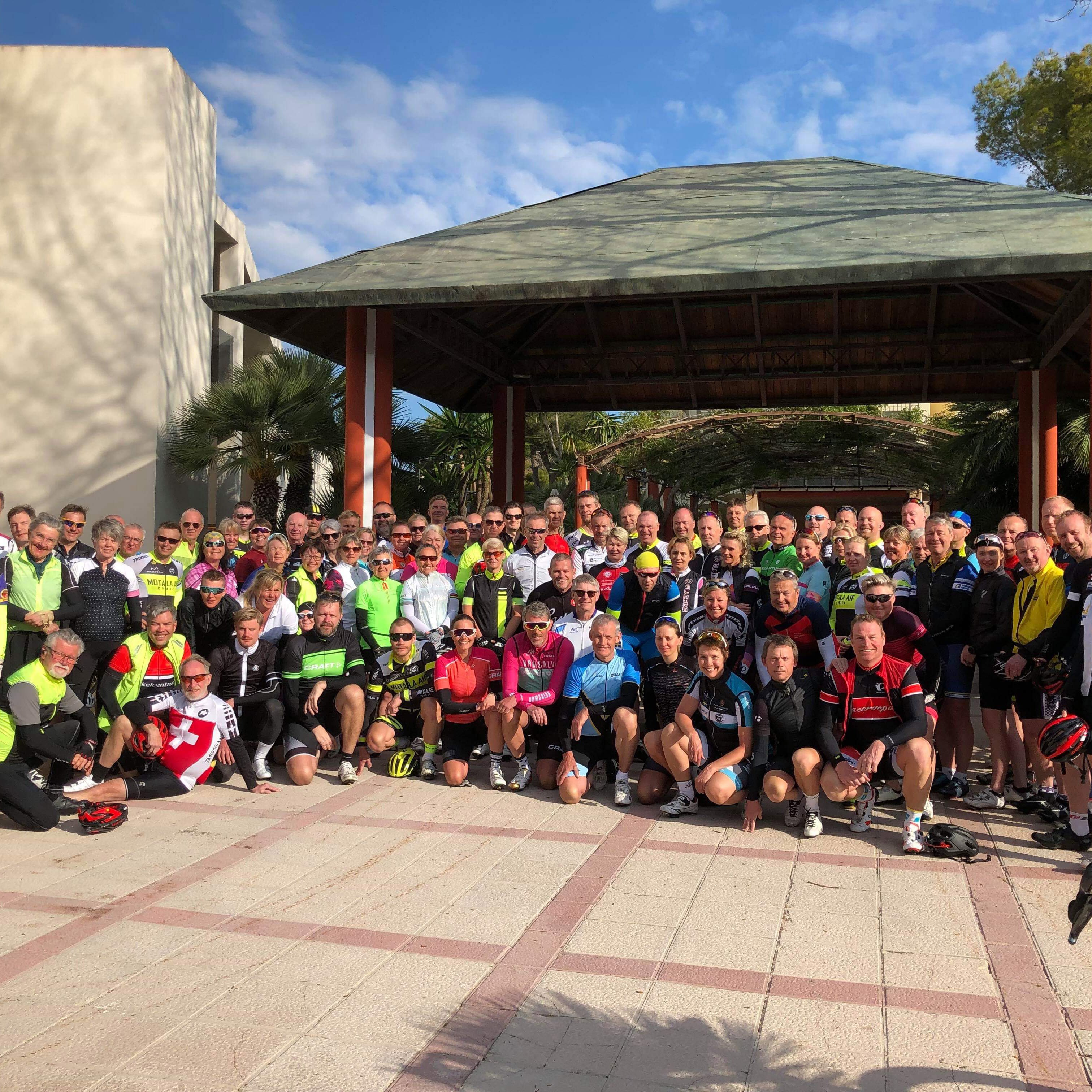 A large group of cyclists in colorful gear poses for a photo outdoors.