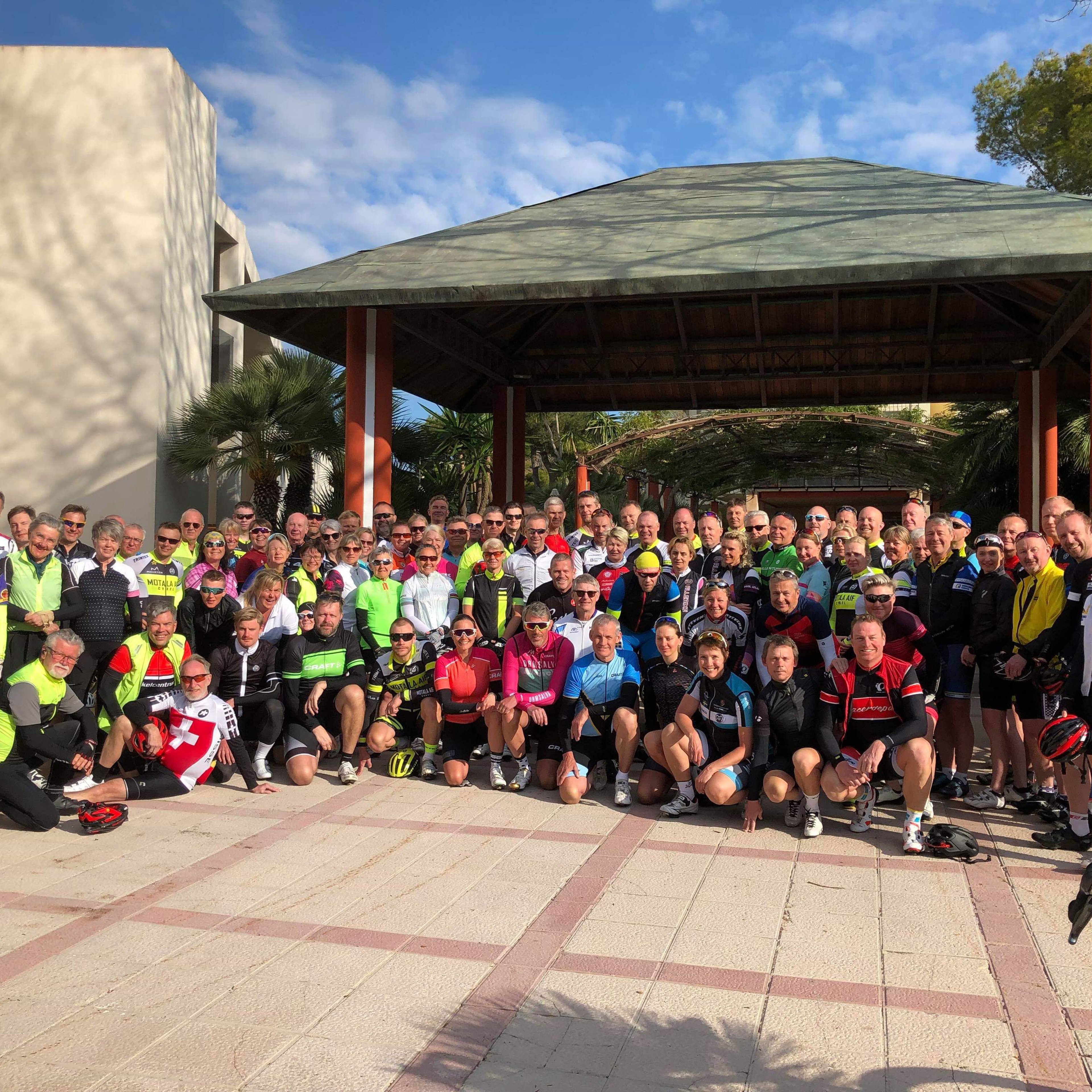 A large group of cyclists in colorful gear poses for a photo outdoors.