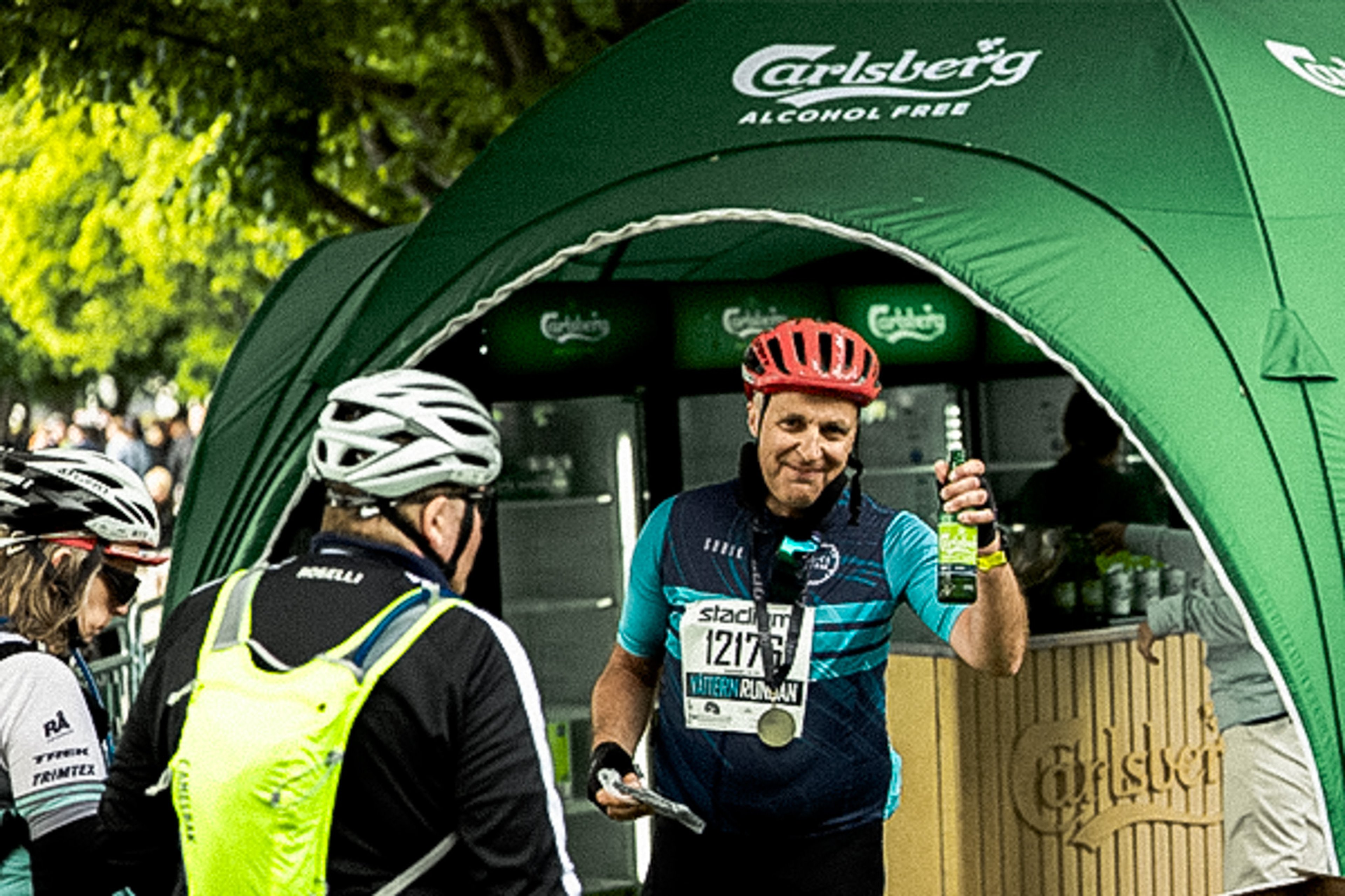 A smiling cyclist in a red helmet and blue jersey holds up a bottle and a medal under a green Carlsberg Alcohol Free tent.