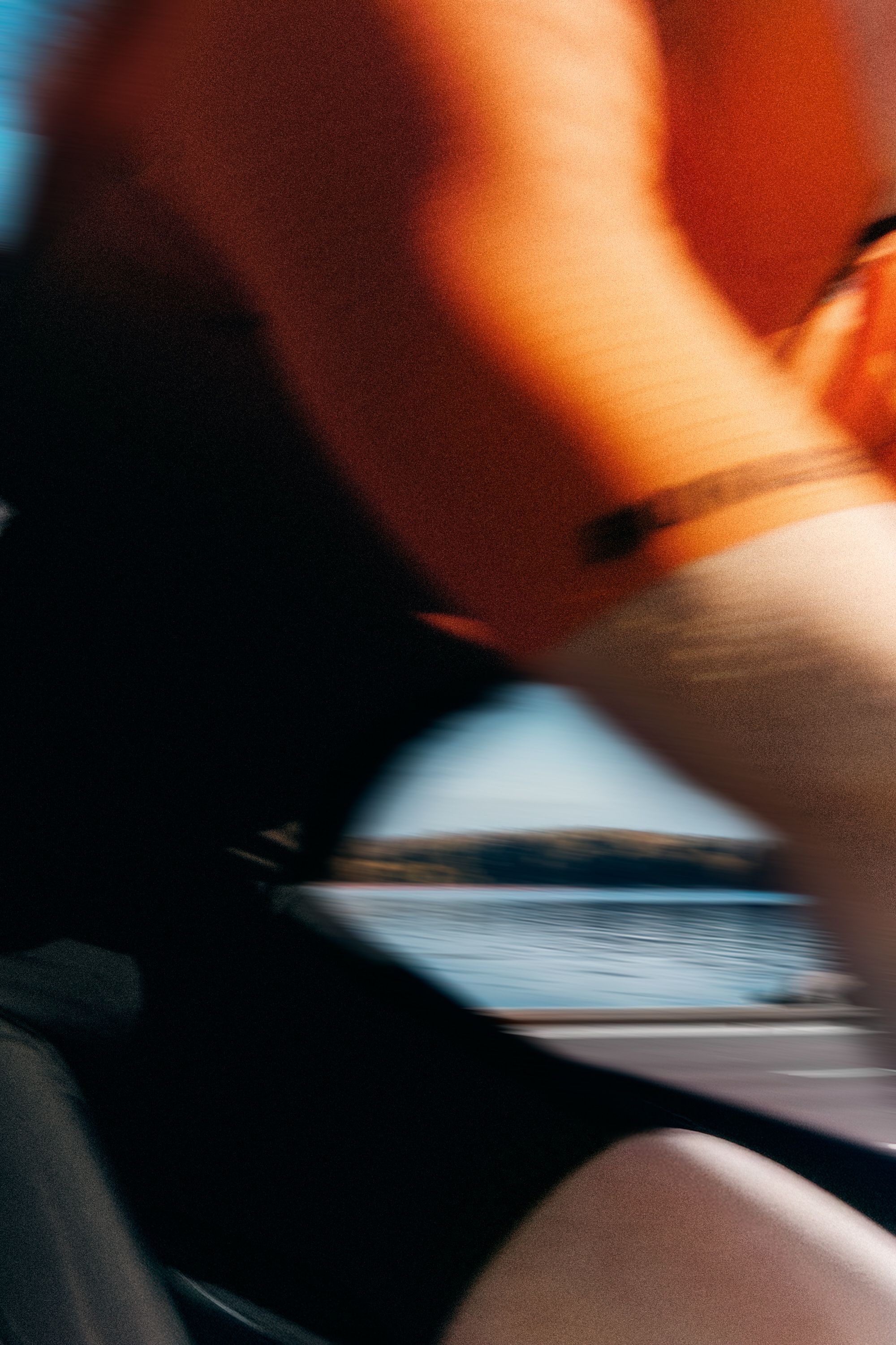 Motion-blurred view from a vehicle, showing an orange-clad leg and a lake with a distant treeline through a window.