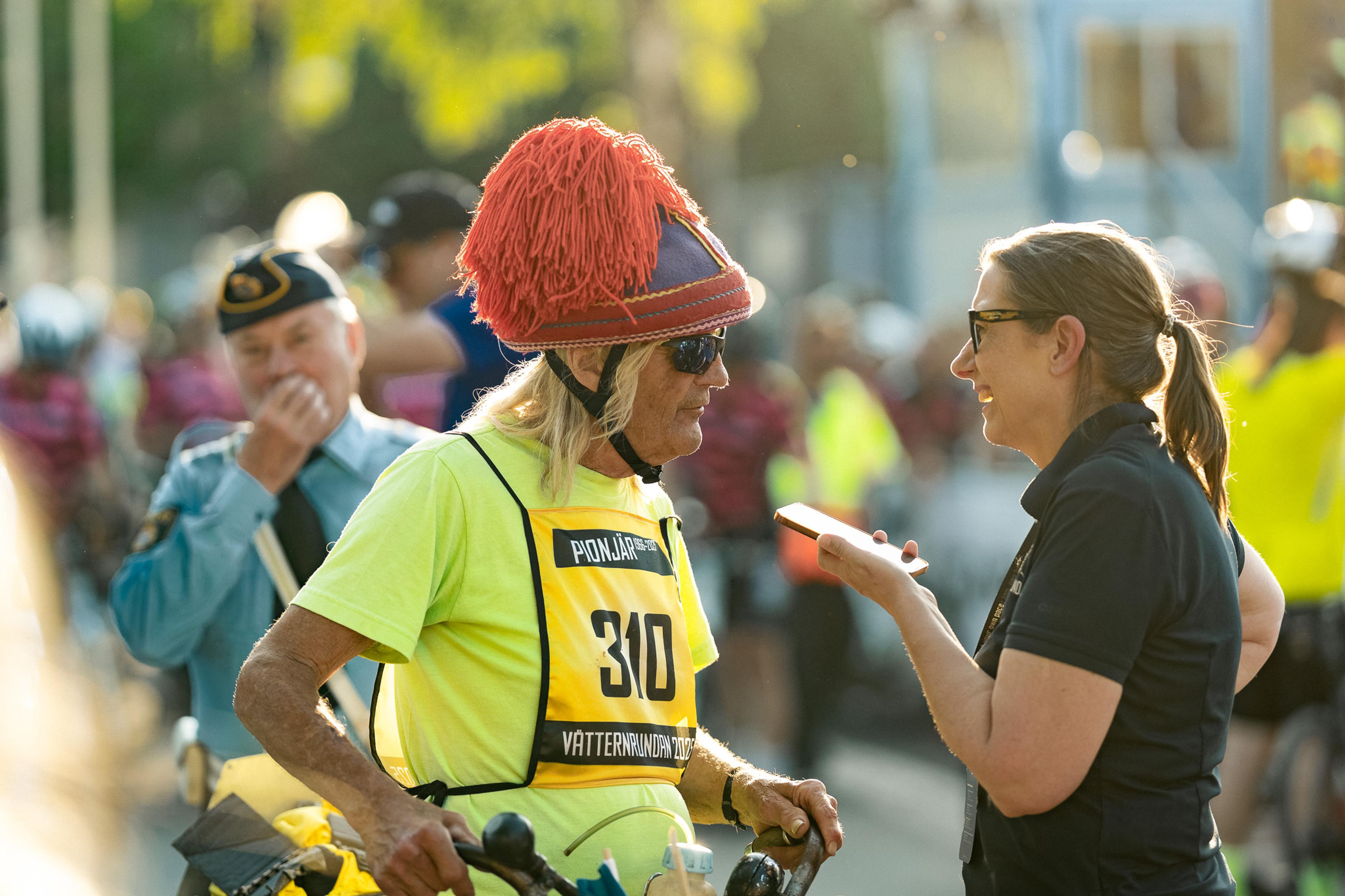 A cyclist in a bright yellow jersey and a red fringed hat talks to a woman holding a phone at an outdoor event.