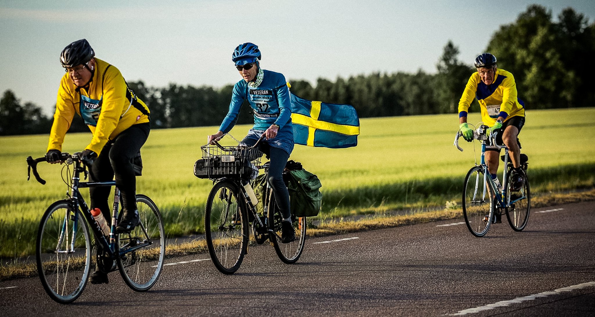 Three cyclists ride on a road beside a green field; one bike carries a Swedish flag.