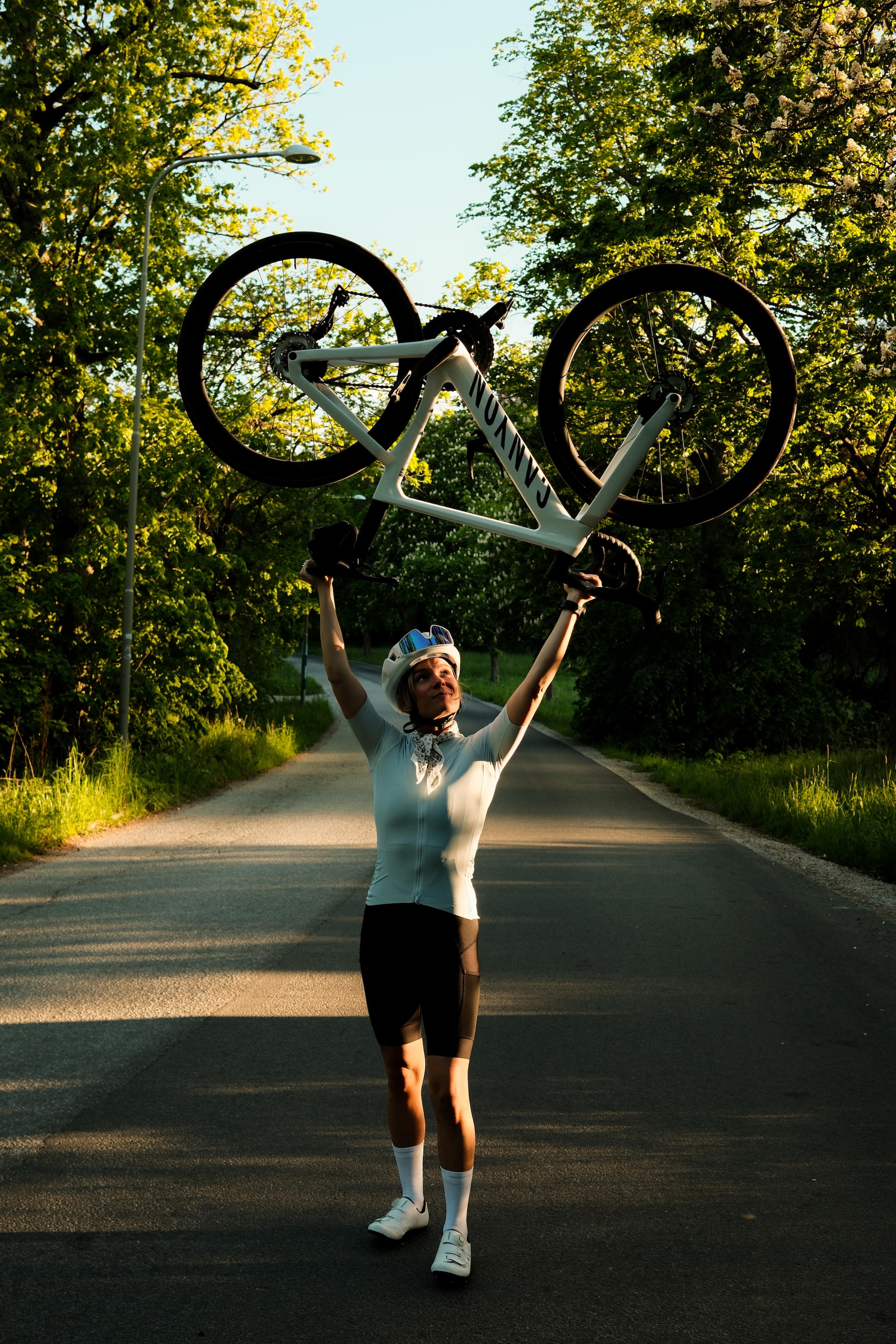 A woman in cycling gear triumphantly lifts a white Canyon road bike above her head on a sunny, tree-lined road.