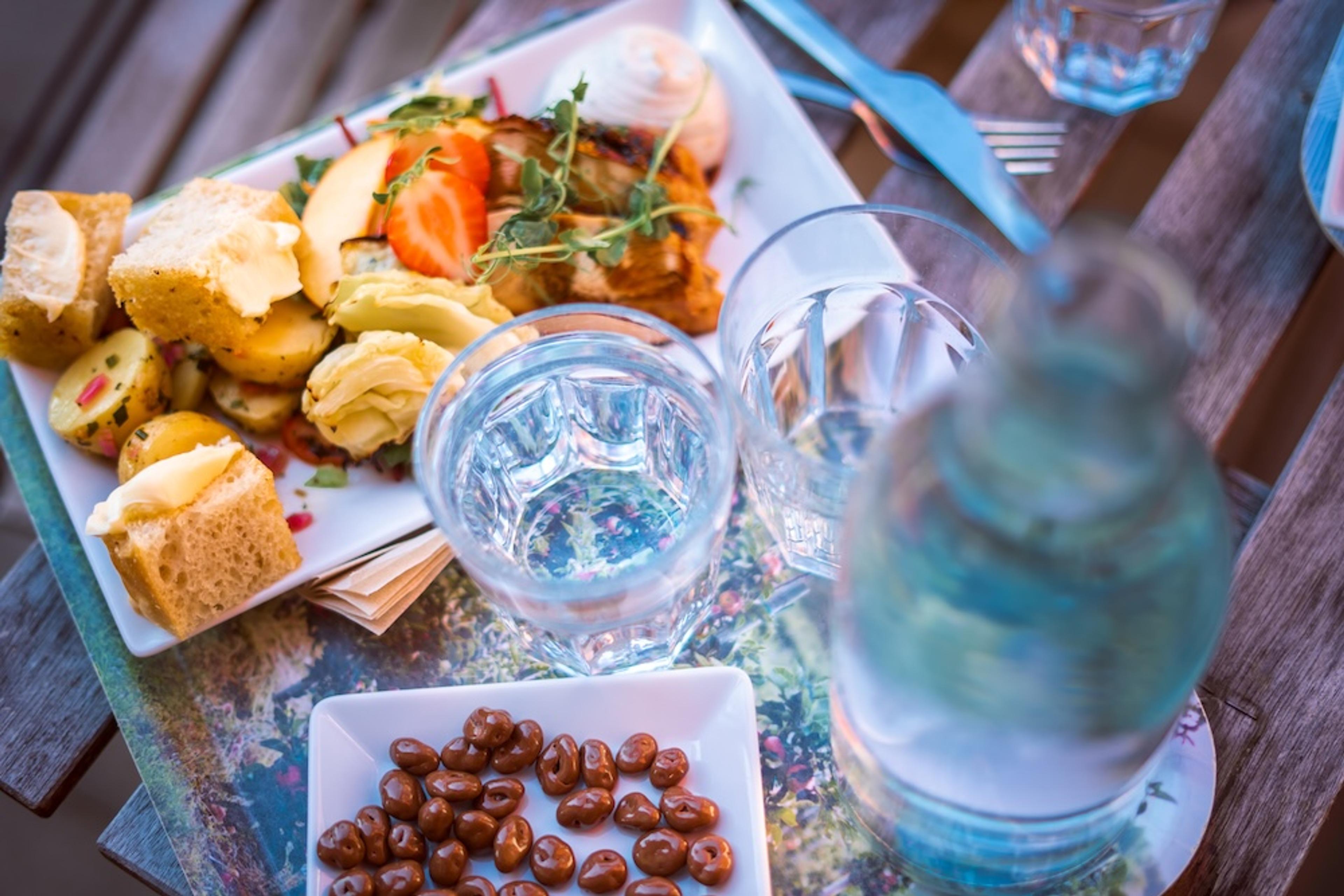 A rustic wooden table set with a plate of bread, potatoes, and salad, along with water glasses, a bottle, and chocolate candies.