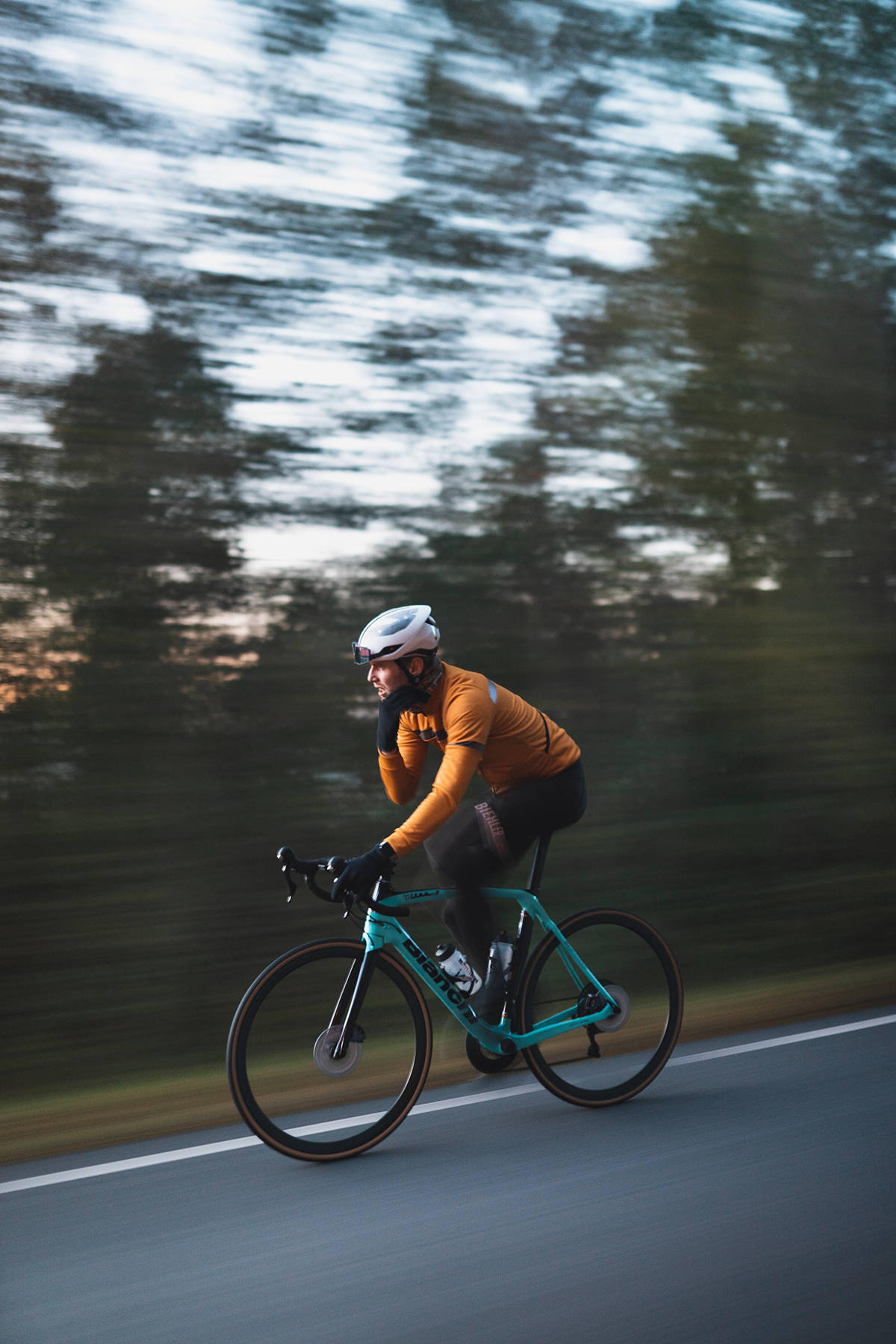 A cyclist in an orange jersey and white helmet rides a teal road bike on a paved road, with a motion-blurred background.